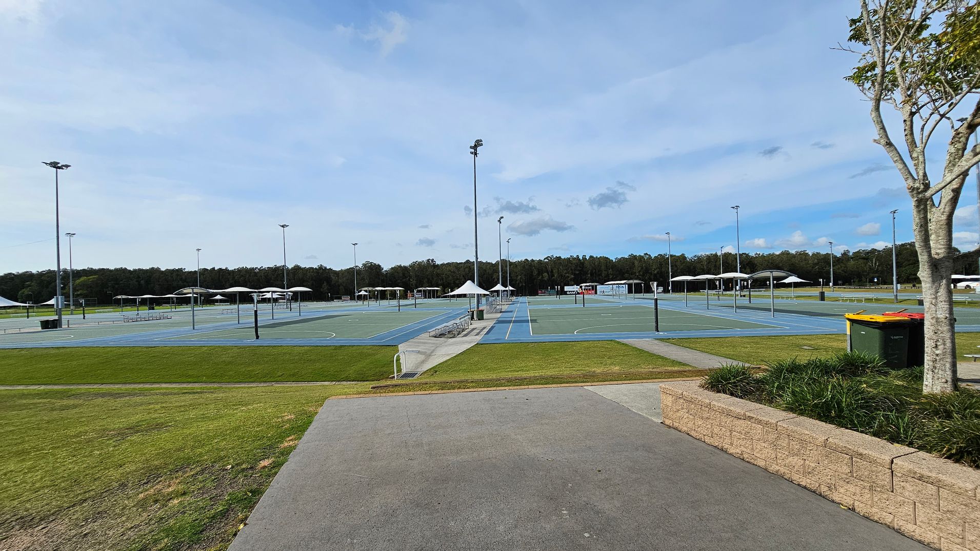 A series of outdoor sports courts with blue surfaces and white lines, surrounded by tall floodlights. In the background, there are several white canopies providing shade. The foreground features a paved path and a grassy area with a low brick wall on the right. A tree and a green rubbish bin with a yellow lid are also on the right. Dense trees form a line in the distant background. A series of outdoor sports courts with blue surfaces and white lines, surrounded by tall floodlights. In the background, there are several white canopies providing shade. The foreground features a paved path and a grassy area with a low brick wall on the right. A tree and a green rubbish bin with a yellow lid are also on the right. Dense trees form a line in the distant background.