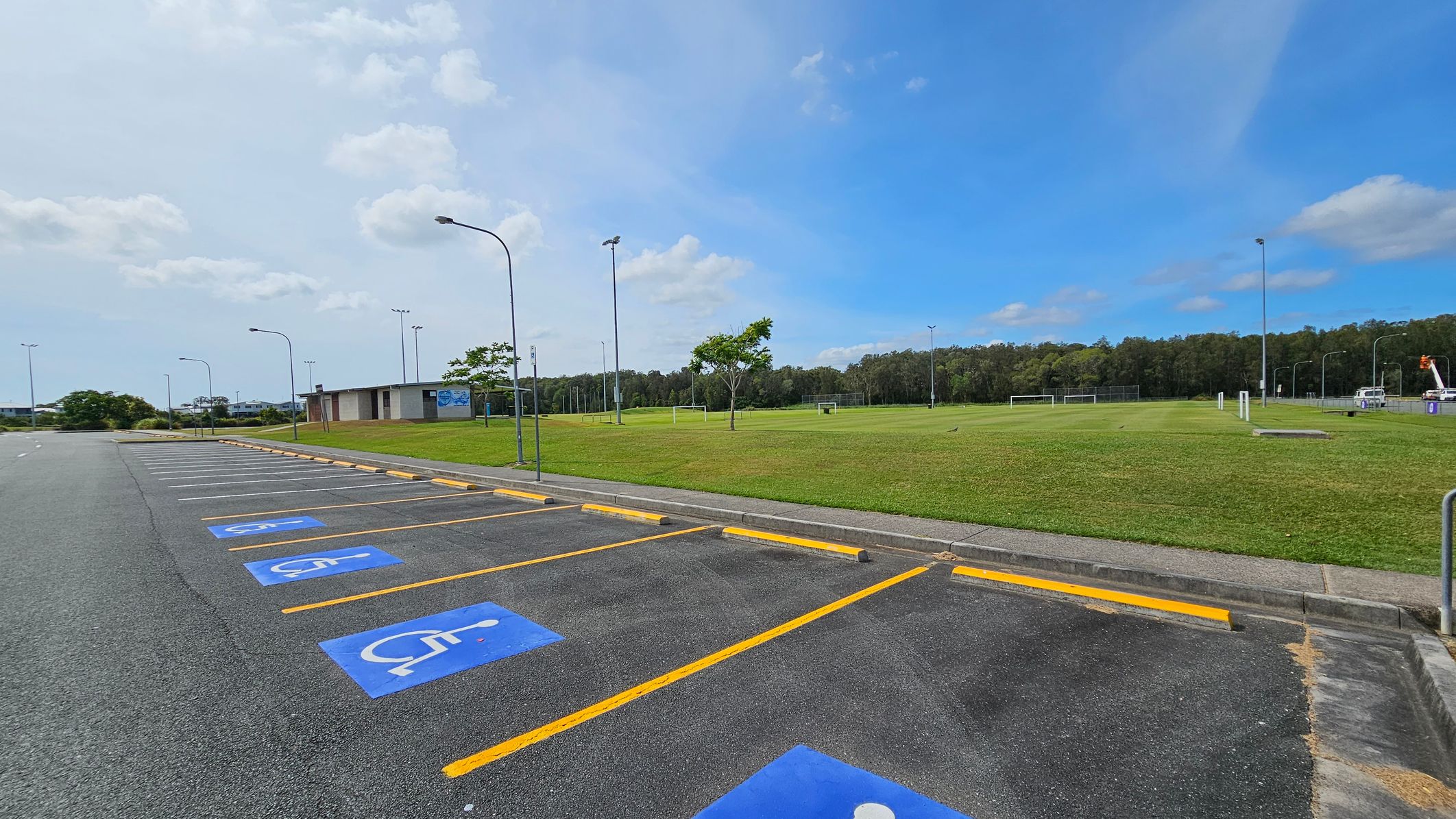 A parking area with several marked disabled parking spaces on the left, featuring blue symbols on the asphalt. Yellow lines separate the spaces. On the right, there is a large grassy field with a few trees and football goals. A small building is visible in the background, along with tall light poles. The area is bordered by a line of trees in the distance. A parking area with several marked disabled parking spaces on the left, featuring blue symbols on the asphalt. Yellow lines separate the spaces. On the right, there is a large grassy field with a few trees and football goals. A small building is visible in the background, along with tall light poles. The area is bordered by a line of trees in the distance.