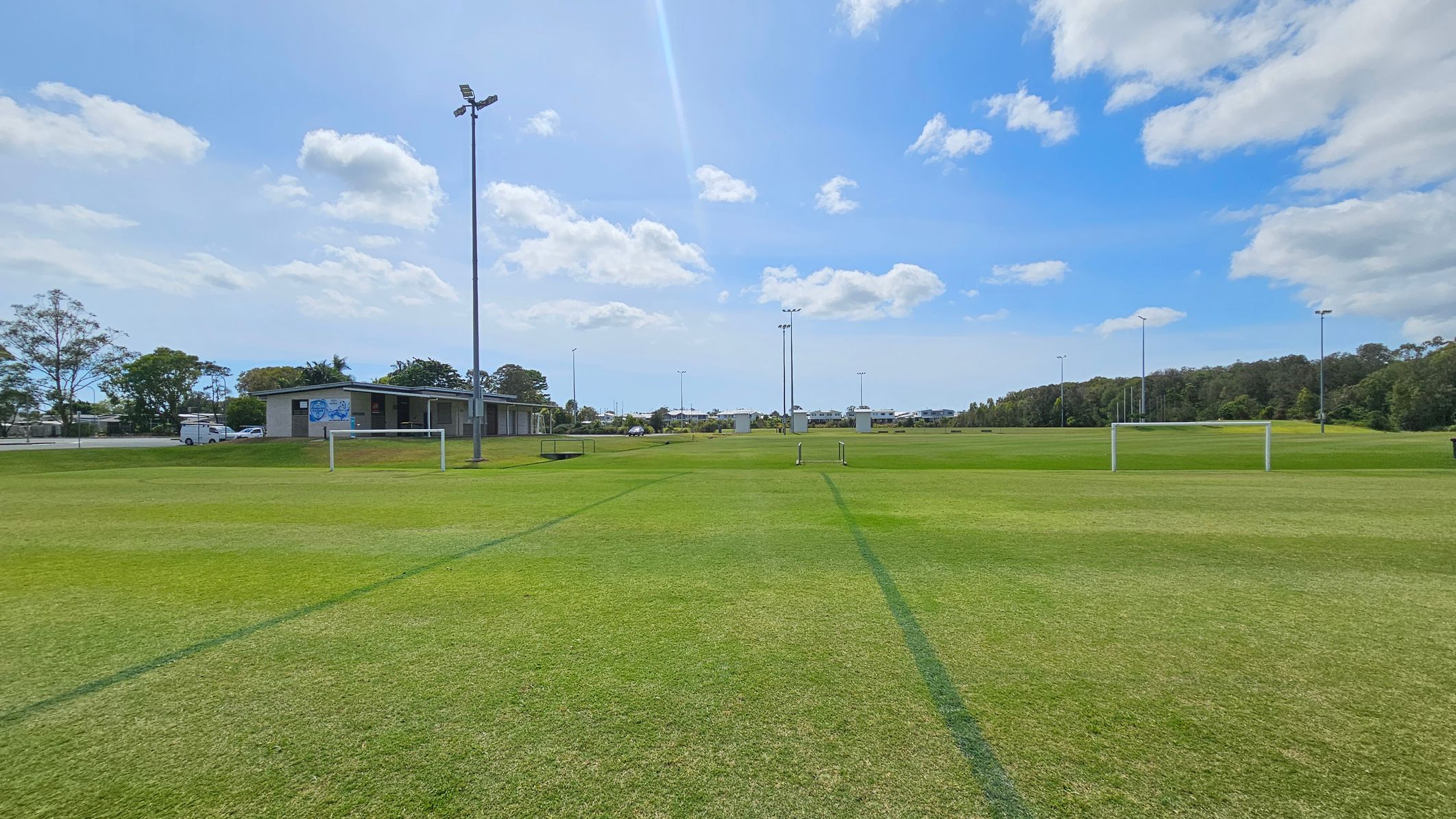 A grassy sports field with goalposts in the foreground. In the background, there is a small building to the left, with a few parked vehicles nearby. Several tall floodlights are scattered across the field. Trees line the horizon on the right side, and a few structures are visible in the distance. The sky is partly cloudy. A grassy sports field with goalposts in the foreground. In the background, there is a small building to the left, with a few parked vehicles nearby. Several tall floodlights are scattered across the field. Trees line the horizon on the right side, and a few structures are visible in the distance. The sky is partly cloudy.