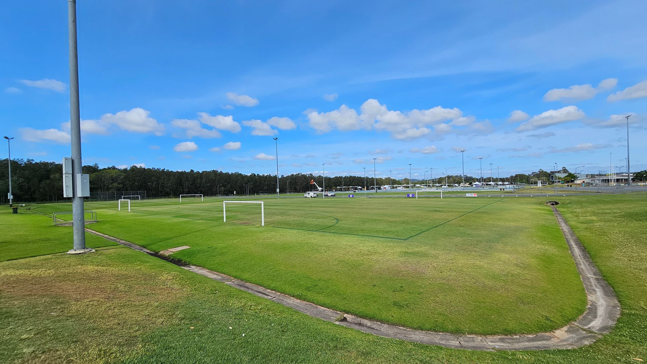 A large grassy sports field with several white goalposts scattered across it. On the left, a tall floodlight pole stands near the edge of the field. In the background, a line of trees borders the field, and a vehicle with a raised platform is visible. To the right, there are more floodlights and a few buildings in the distance. The ground is a mix of green and brown patches, with a narrow concrete drainage channel running through it. A large grassy sports field with several white goalposts scattered across it. On the left, a tall floodlight pole stands near the edge of the field. In the background, a line of trees borders the field, and a vehicle with a raised platform is visible. To the right, there are more floodlights and a few buildings in the distance. The ground is a mix of green and brown patches, with a narrow concrete drainage channel running through it.