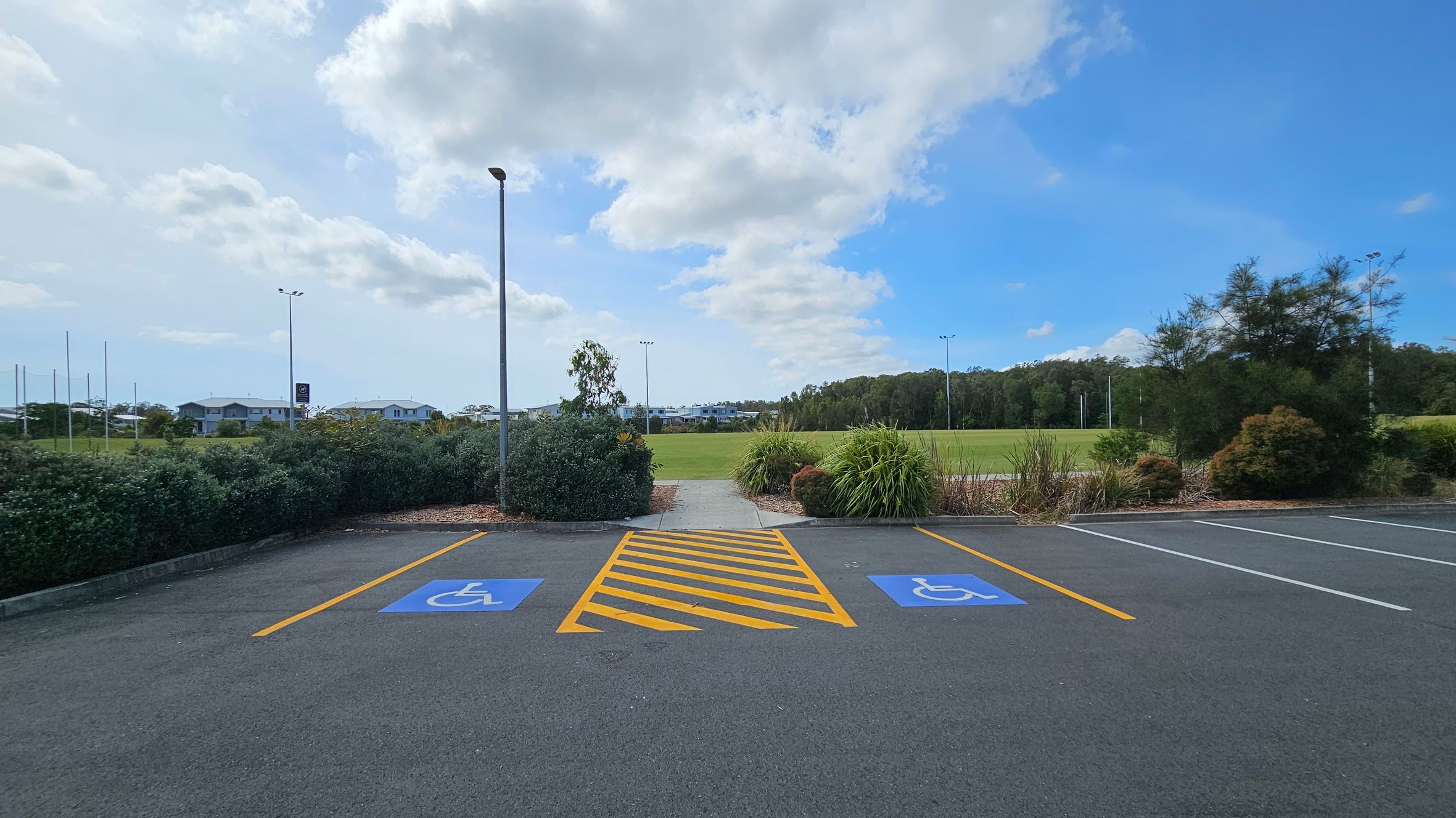 Two parking spaces with blue disabled symbols are marked on the asphalt, separated by yellow diagonal lines. A path leads from the parking area through low bushes and greenery towards a grassy field in the background. A tall lamp post stands near the path. In the distance, there are houses and trees lining the horizon. Two parking spaces with blue disabled symbols are marked on the asphalt, separated by yellow diagonal lines. A path leads from the parking area through low bushes and greenery towards a grassy field in the background. A tall lamp post stands near the path. In the distance, there are houses and trees lining the horizon.