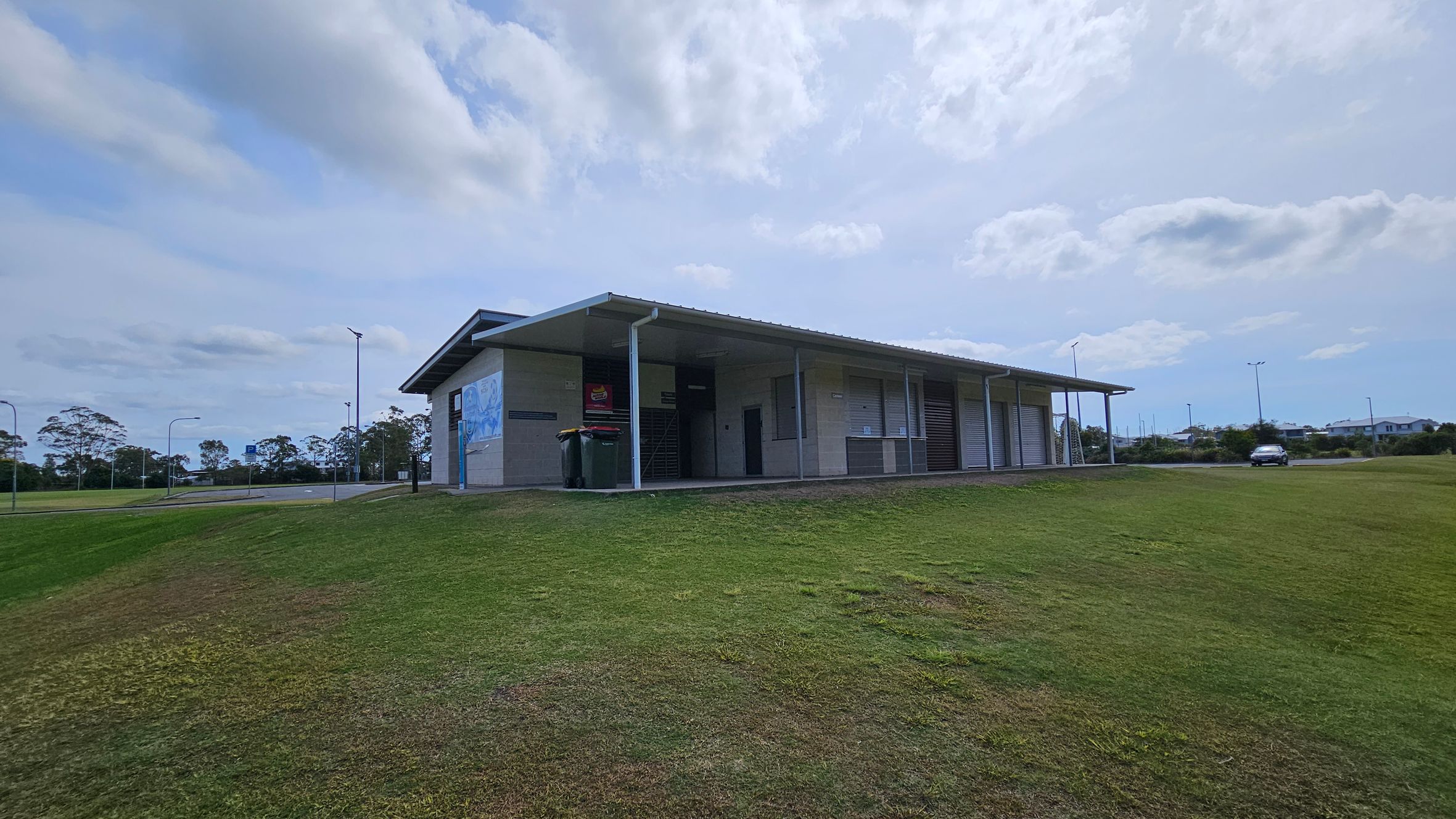 A single-storey building with a sloped roof is situated on a grassy hill. The structure has a covered walkway with several roller doors and a mural on the left wall. Two bins are placed near the entrance. In the background, there are trees and a few streetlights. A car is parked on the right side, near a road that runs alongside the building. A single-storey building with a sloped roof is situated on a grassy hill. The structure has a covered walkway with several roller doors and a mural on the left wall. Two bins are placed near the entrance. In the background, there are trees and a few streetlights. A car is parked on the right side, near a road that runs alongside the building.