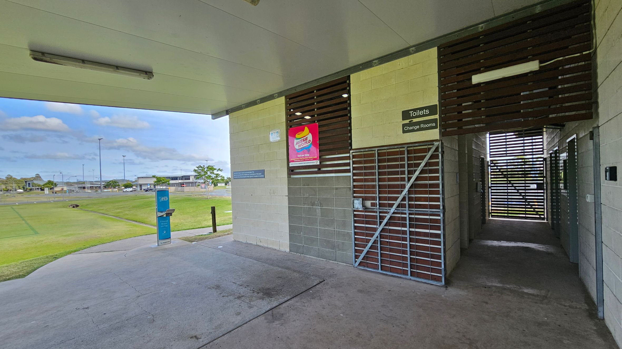A sports facility with a covered area. On the right, there is a gated entrance to toilets and change rooms, with a sign above. A poster is displayed on the wall. The floor is concrete. On the left, an open grassy field is visible, with a water fountain in the foreground. In the background, there are buildings and a car park. The ceiling is flat with a light fixture. A sports facility with a covered area. On the right, there is a gated entrance to toilets and change rooms, with a sign above. A poster is displayed on the wall. The floor is concrete. On the left, an open grassy field is visible, with a water fountain in the foreground. In the background, there are buildings and a car park. The ceiling is flat with a light fixture.