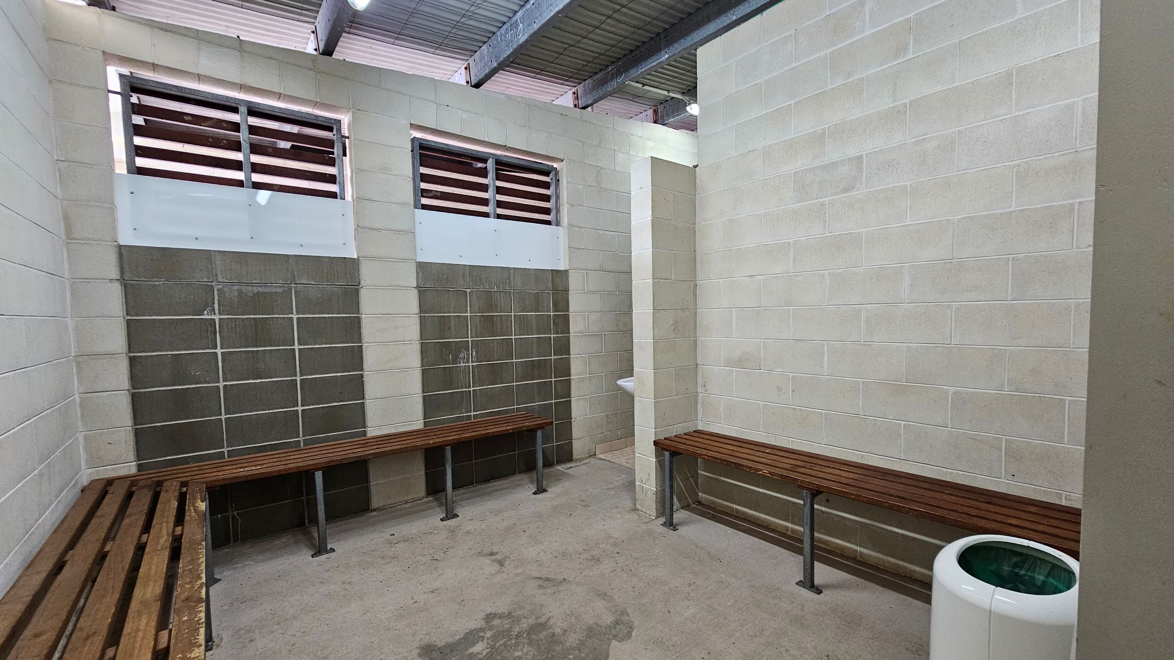 A room with concrete block walls and a corrugated metal ceiling. On the left and right, there are wooden benches attached to the walls. Two small windows with horizontal slats are on the left wall. In the corner, a small white sink is mounted on the wall. The floor is concrete with visible stains. A white cylindrical bin with a green interior is on the right side. A room with concrete block walls and a corrugated metal ceiling. On the left and right, there are wooden benches attached to the walls. Two small windows with horizontal slats are on the left wall. In the corner, a small white sink is mounted on the wall. The floor is concrete with visible stains. A white cylindrical bin with a green interior is on the right side.