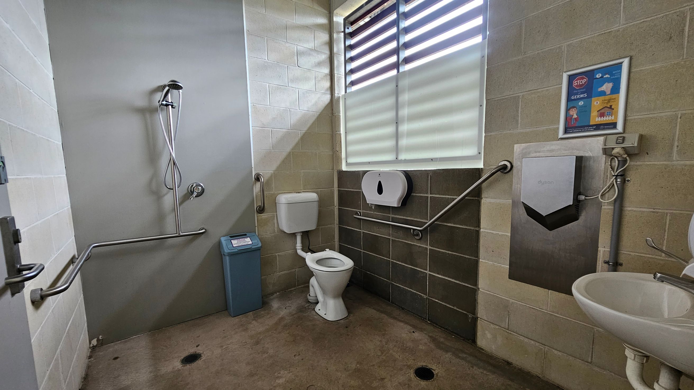 A public bathroom with a concrete floor and tiled walls. On the left, there's a shower with a handheld showerhead and a metal grab bar. In the centre, a toilet with a cistern is positioned next to a blue bin. On the right, a wall-mounted hand dryer and a paper towel dispenser are above a sink. A poster about hygiene is on the wall. The window has horizontal slats allowing light in. A public bathroom with a concrete floor and tiled walls. On the left, there's a shower with a handheld showerhead and a metal grab bar. In the centre, a toilet with a cistern is positioned next to a blue bin. On the right, a wall-mounted hand dryer and a paper towel dispenser are above a sink. A poster about hygiene is on the wall. The window has horizontal slats allowing light in.