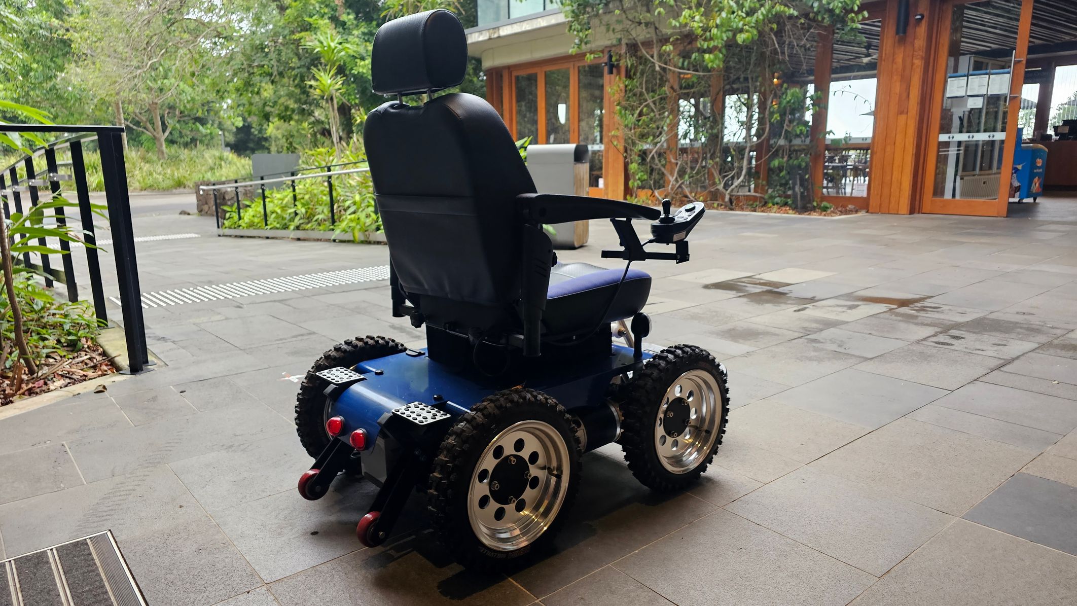 A motorised wheelchair with large, rugged tyres is positioned on a tiled floor. The wheelchair has a black seat with armrests and a joystick control on the right armrest. In the background, there is a building with wooden and glass elements, surrounded by greenery and trees. A metal railing is visible on the left, and the area appears to be an outdoor patio or walkway.