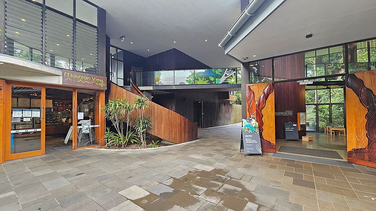 On the left, there is a café with wooden framed glass doors and a sign reading "Mountain View Café". Inside, a counter with menus is visible. In the centre, a staircase with wooden panelling leads to an upper level with glass railings. On the right, large wooden doors with carved designs open into a space with a sign for entry and a donation box. The floor is paved with multicoloured tiles, and the ceiling is smooth with recessed lighting.