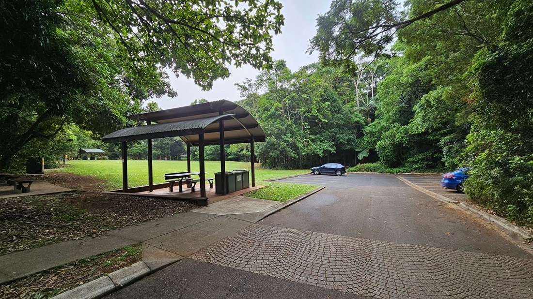 A park scene with a sheltered picnic area on the left, featuring a table and benches under a curved roof. The ground is covered with leaves and grass. In the background, a grassy field is visible, bordered by dense green trees. On the right, there is a paved parking area with two cars, one black and one blue. The foreground shows a textured pavement leading to the parking area.
