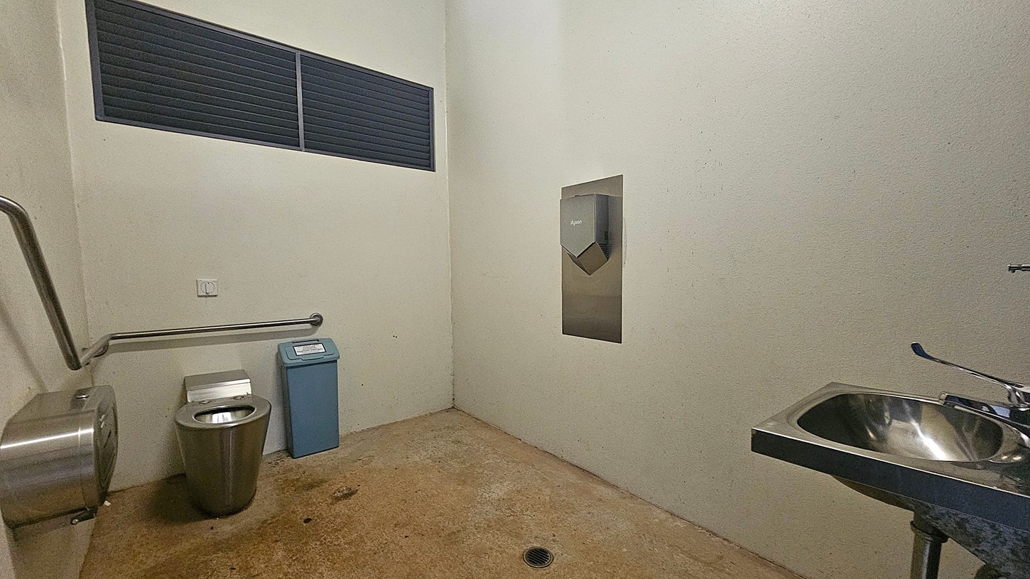 A public toilet with a stainless steel toilet on the left, accompanied by a toilet paper dispenser and a grab rail. Next to it is a blue sanitary bin. On the right, a stainless steel sink with a tap is visible. A hand dryer is mounted on the wall above the sink. The floor is concrete with visible stains, and the walls are light-coloured. A black vent is located high on the left wall.