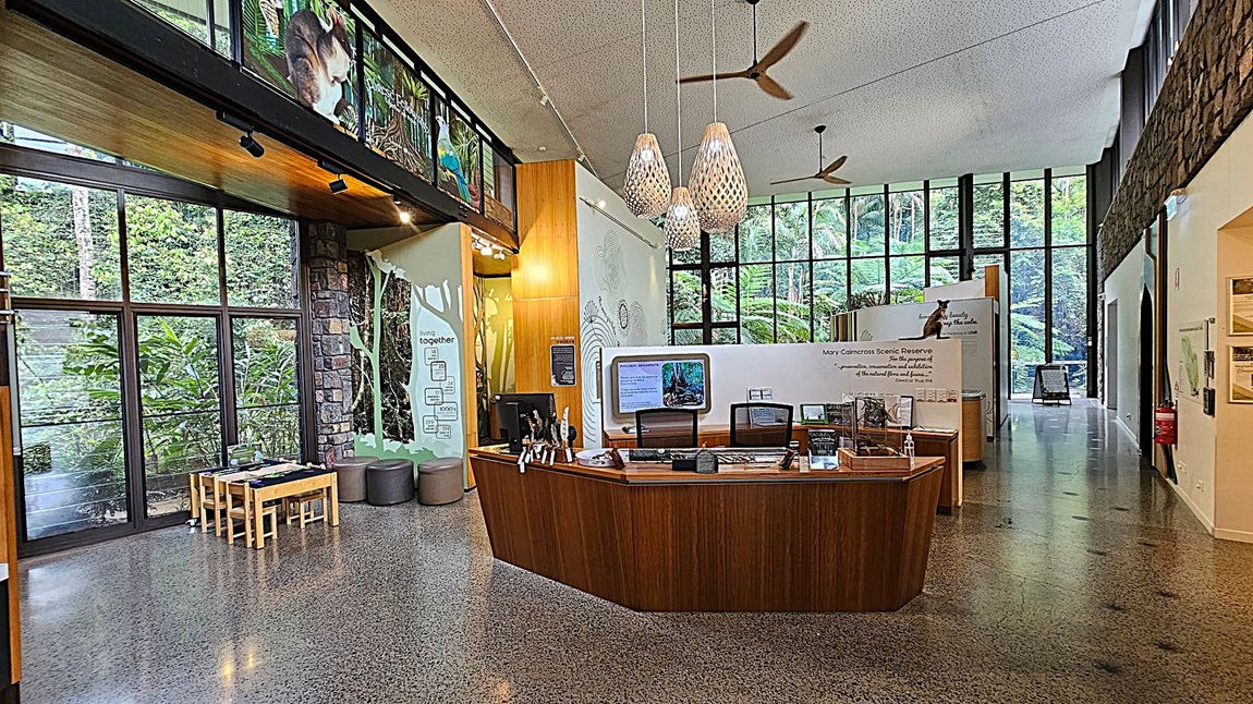 A spacious interior with a polished concrete floor and high ceiling. On the left, large windows reveal lush greenery outside. Below the windows, a small wooden table with chairs is set up. In the centre, a wooden reception desk displays informational materials. Above, three pendant lights hang from the ceiling. The right side features more large windows and a stone wall, with informational displays about the Mary Cairncross Scenic Reserve.