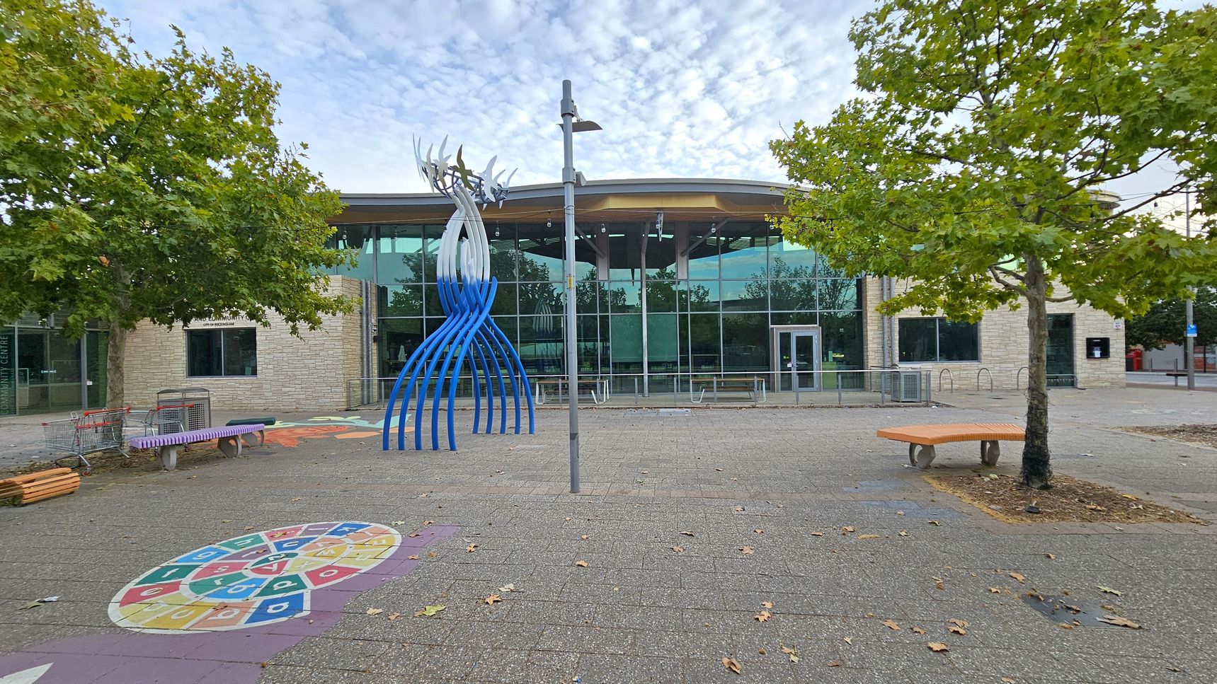 A modern building with large glass windows is in the background. In the foreground, there is a blue and white abstract sculpture. To the left, a tree with green leaves and a purple bench are visible, along with some shopping trolleys. On the ground, there is a colourful circular pattern with numbers. To the right, another tree and an orange bench are present. A lamppost stands in the centre of the paved area, which is scattered with fallen leaves.