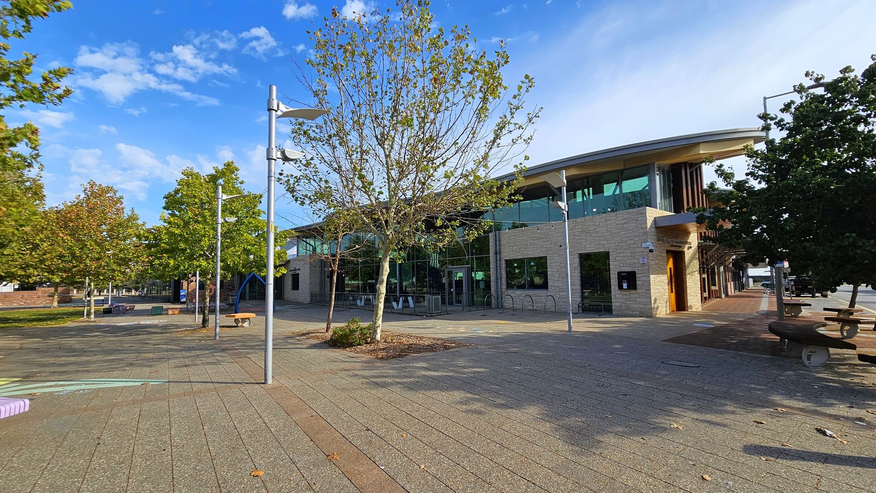 A modern building with large glass windows and a light-coloured brick facade is on the right. In front, there are several trees with sparse leaves. The foreground features a paved area with a few benches and street lamps. On the left, more trees line the area, and there are some scattered benches and play equipment. The scene is well-lit, suggesting daytime.