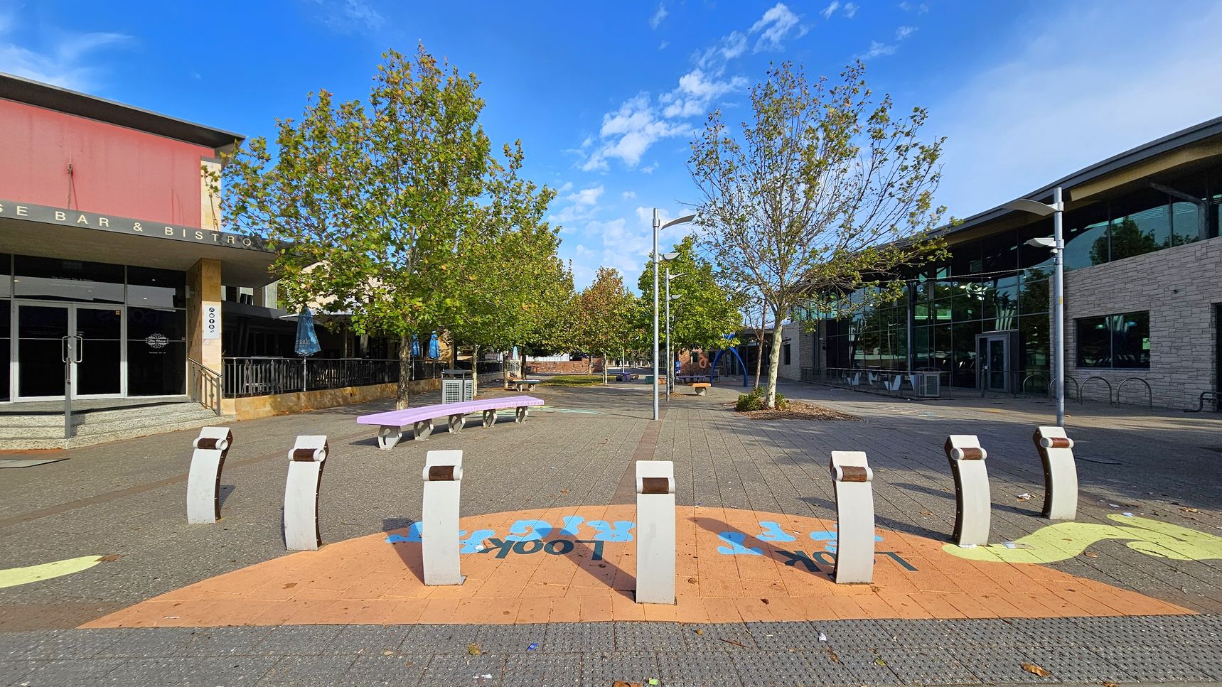 A paved outdoor area with several white bollards in the foreground. On the left, there's a building with a sign reading "BAR & BISTRO" and a pink facade. In the centre, trees line the pathway, with benches and street lamps visible. On the right, a modern building with large windows and a stone exterior. The ground features colourful painted designs, and the sky is clear with a few clouds.