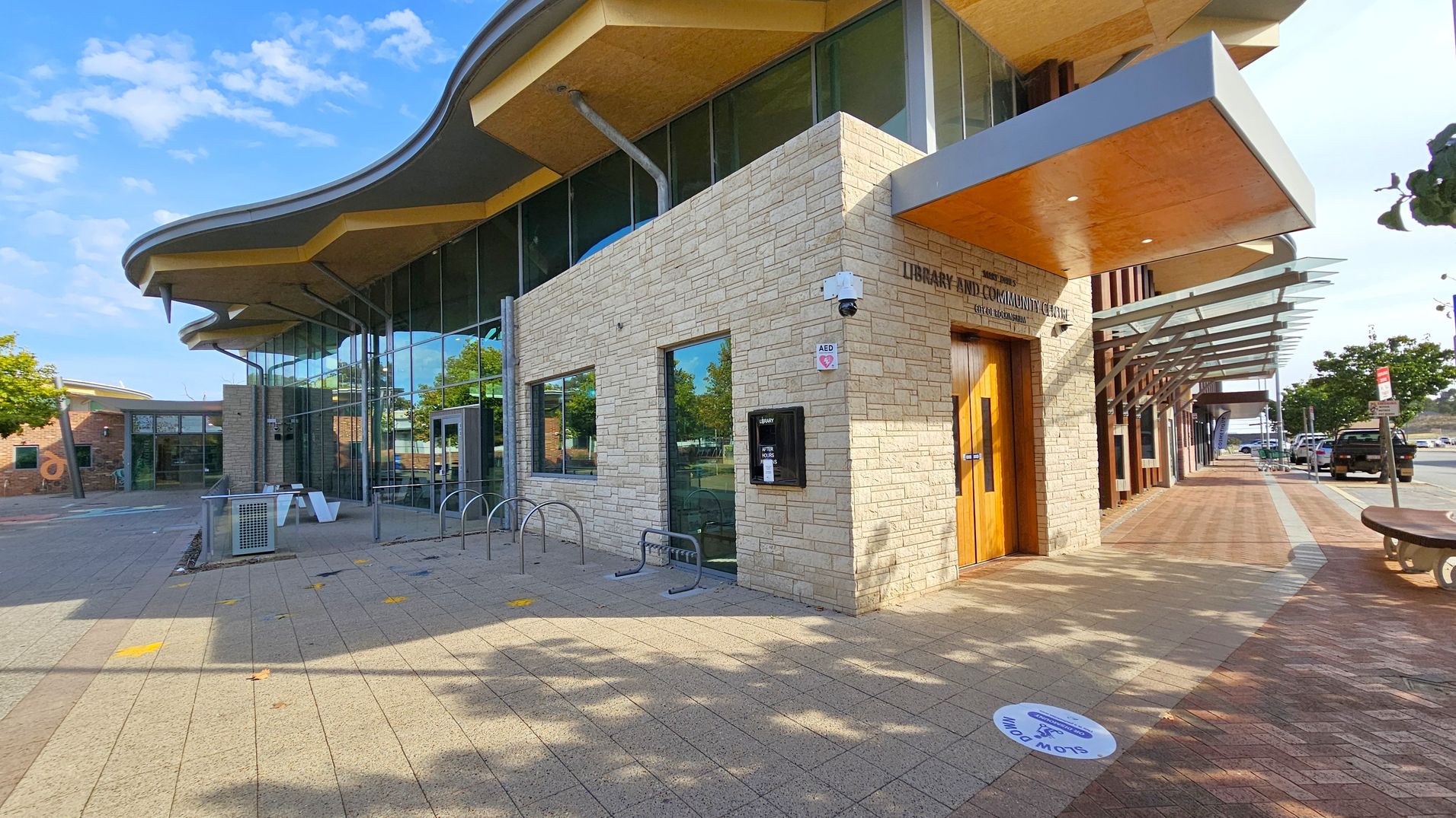 A modern building with a wavy roofline and large glass windows. The left side features a stone facade with a bike rack and a bench nearby. The entrance has a wooden door with a small canopy above it. On the right, a covered walkway with a glass and metal awning extends along the pavement. The ground is paved with a mix of light and dark tiles, and there are trees and parked cars in the background.