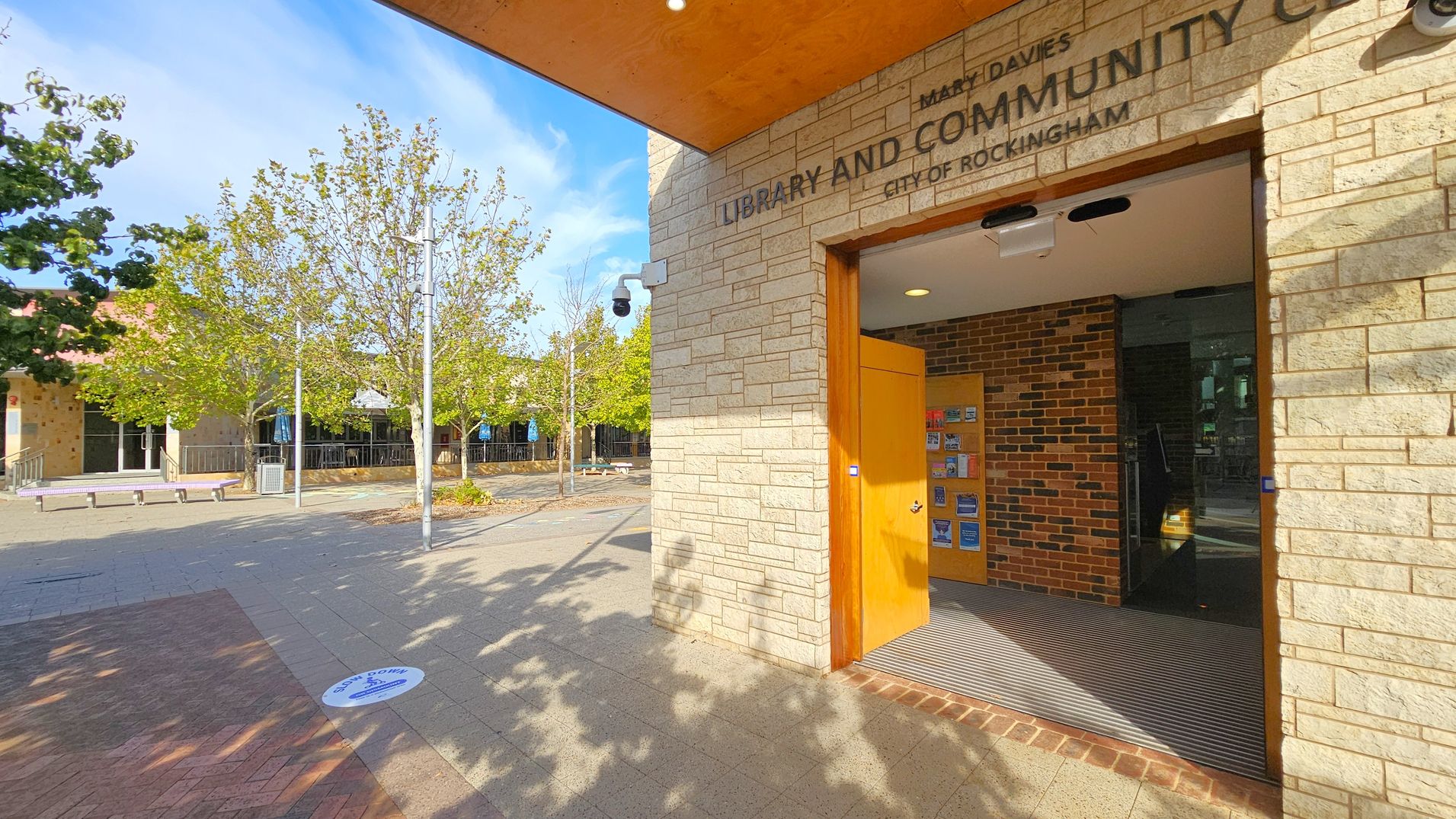 Entrance to a library and community centre with a stone facade on the right. The door is open, revealing a brick interior wall and a display of brochures. A security camera is mounted above. On the left, a paved area with trees and benches is visible. The ground has a circular social distancing marker. The ceiling above the entrance is orange.