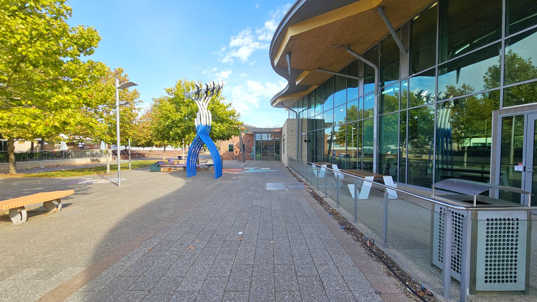 On the left, there are several trees with green leaves and a few benches along a paved walkway. A blue and white abstract sculpture stands prominently. On the right, a modern building with large glass windows reflects the surroundings. The building has a curved roof and a metal railing with a bin nearby. The ground is paved with grey tiles, and there are a few scattered leaves.