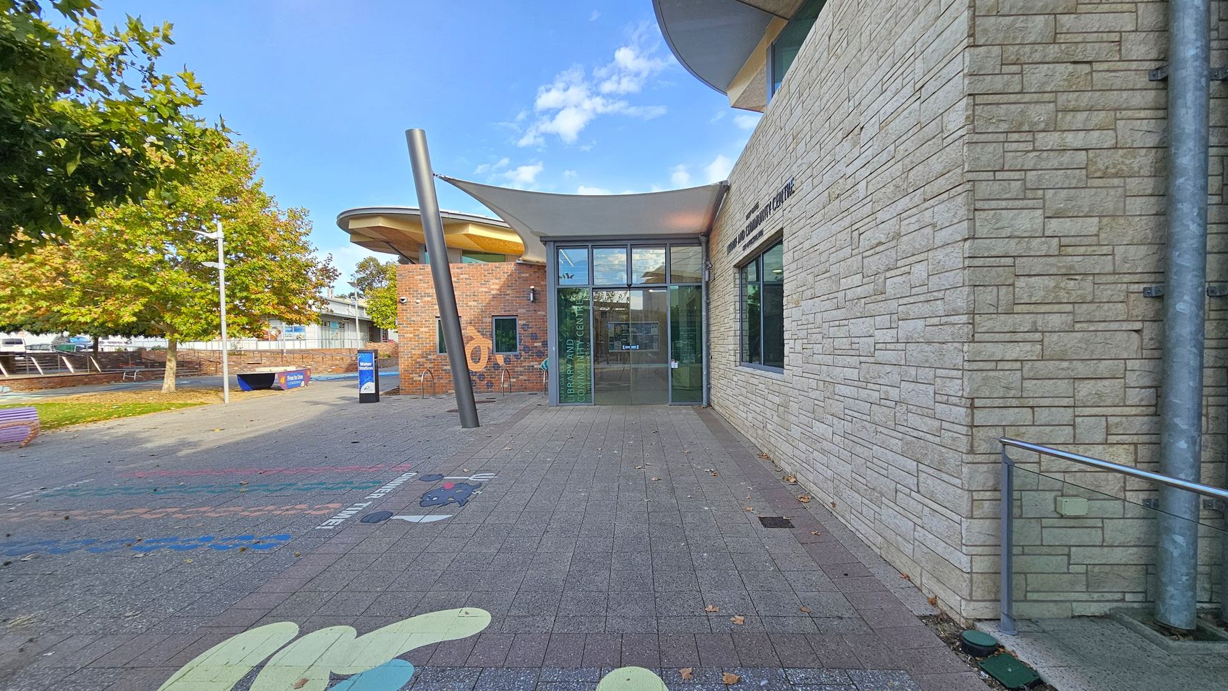 A modern building with a stone facade on the right and a glass entrance in the centre. The ground is paved with colourful patterns. A large, tilted metal pole stands near the entrance. On the left, there are trees with green and yellow leaves, and a brick wall with a circular roof structure. A blue sign and a bench are visible in the background.