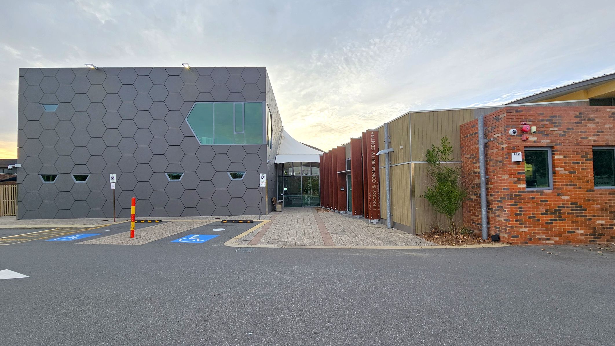 A modern building with a hexagonal-patterned facade on the left, featuring small and large windows. In the centre, a glass entrance with a canopy leads to the interior. On the right, a combination of wooden and brick walls, with a sign indicating a library and community centre. The foreground has a paved path and parking spaces, including a disabled parking spot marked in blue. A small tree is planted near the brick wall.