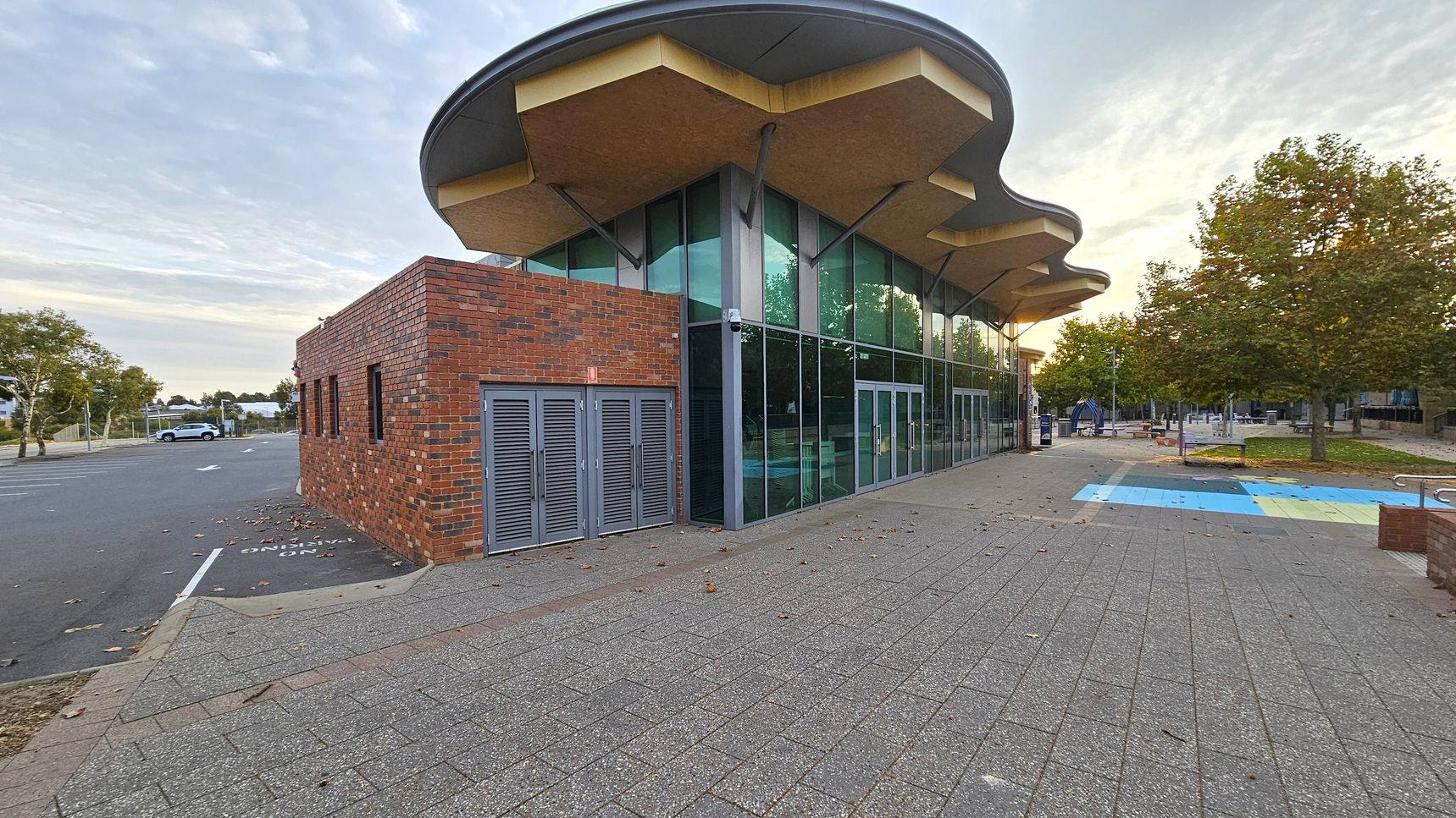 A modern building with a distinctive wavy roof and large glass windows. On the left, there's a brick section with grey double doors. The foreground features a paved area with scattered leaves. To the right, a colourful path leads to a small park with trees and benches. A car is parked on the street to the left, and the background shows more trees and a clear sky.