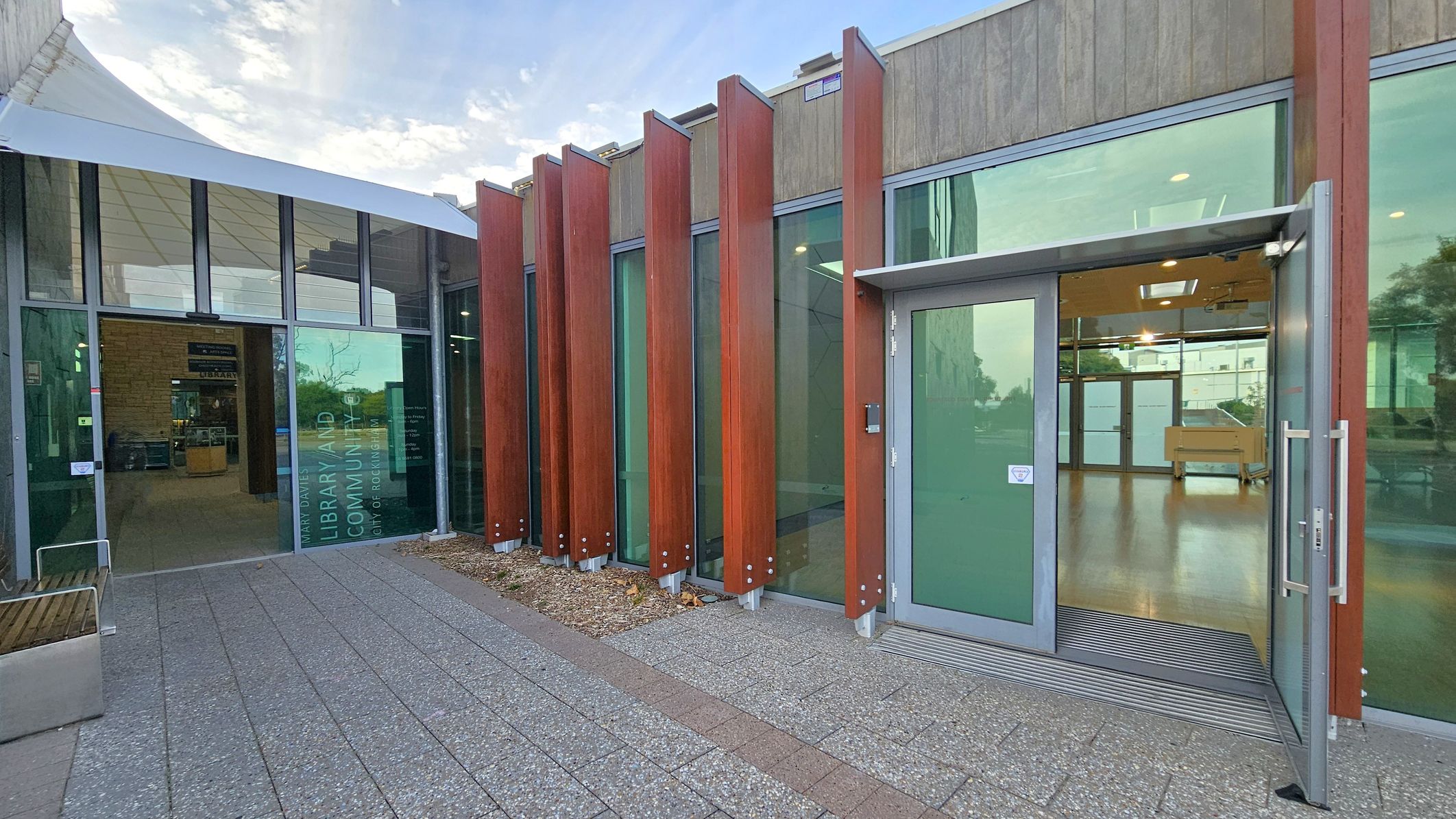 On the left, there is a glass entrance with a sign that reads "Library and Community." Inside, a reception area is visible. The right side features a series of tall, vertical wooden panels in front of large glass windows. An open glass door leads into a room with wooden flooring. The ground is paved with grey and red tiles, and a white canopy extends over the entrance on the left.