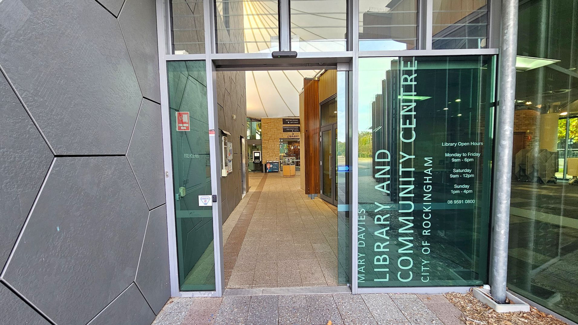Entrance to a library and community centre. On the left, there is a wall with large hexagonal tiles. The entrance door is glass, with text displaying the library's name and opening hours. Inside, a tiled floor leads to a reception area with a wooden panelled wall and a sign for the library. On the right, large windows allow natural light to enter, and there are reflections of trees outside. The ceiling is high with a white, tent-like structure.