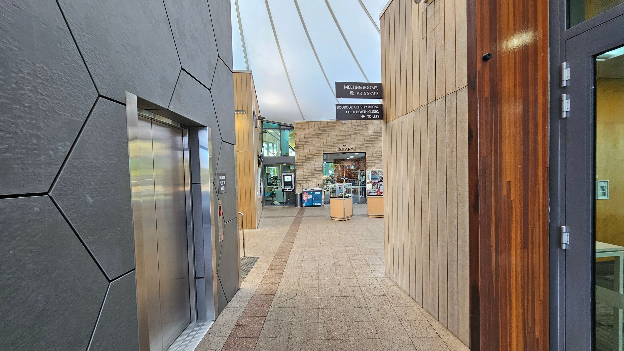 A modern indoor hallway with a geometric-patterned wall on the left featuring a lift. The floor is tiled with a central strip leading towards a library entrance in the background. On the right, there is a wooden-panelled wall and a glass door. Overhead, a curved ceiling with white panels and metal beams. Signs above indicate directions to meeting rooms, an arts space, a child health clinic, and toilets.