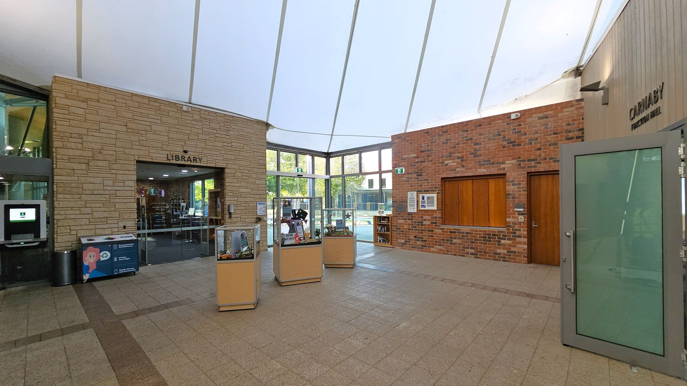 A spacious interior with a high, white ceiling. On the left, a stone wall with a sign for the library and a glass entrance. Below, a digital kiosk and a recycling bin. In the centre, three display cases with various items. The right side features a brick wall with a wooden door and a window. A glass door is open, leading to a room labelled "Carnaby Function Hall." The floor is tiled in a light brown pattern. Large windows in the background allow natural light to enter.