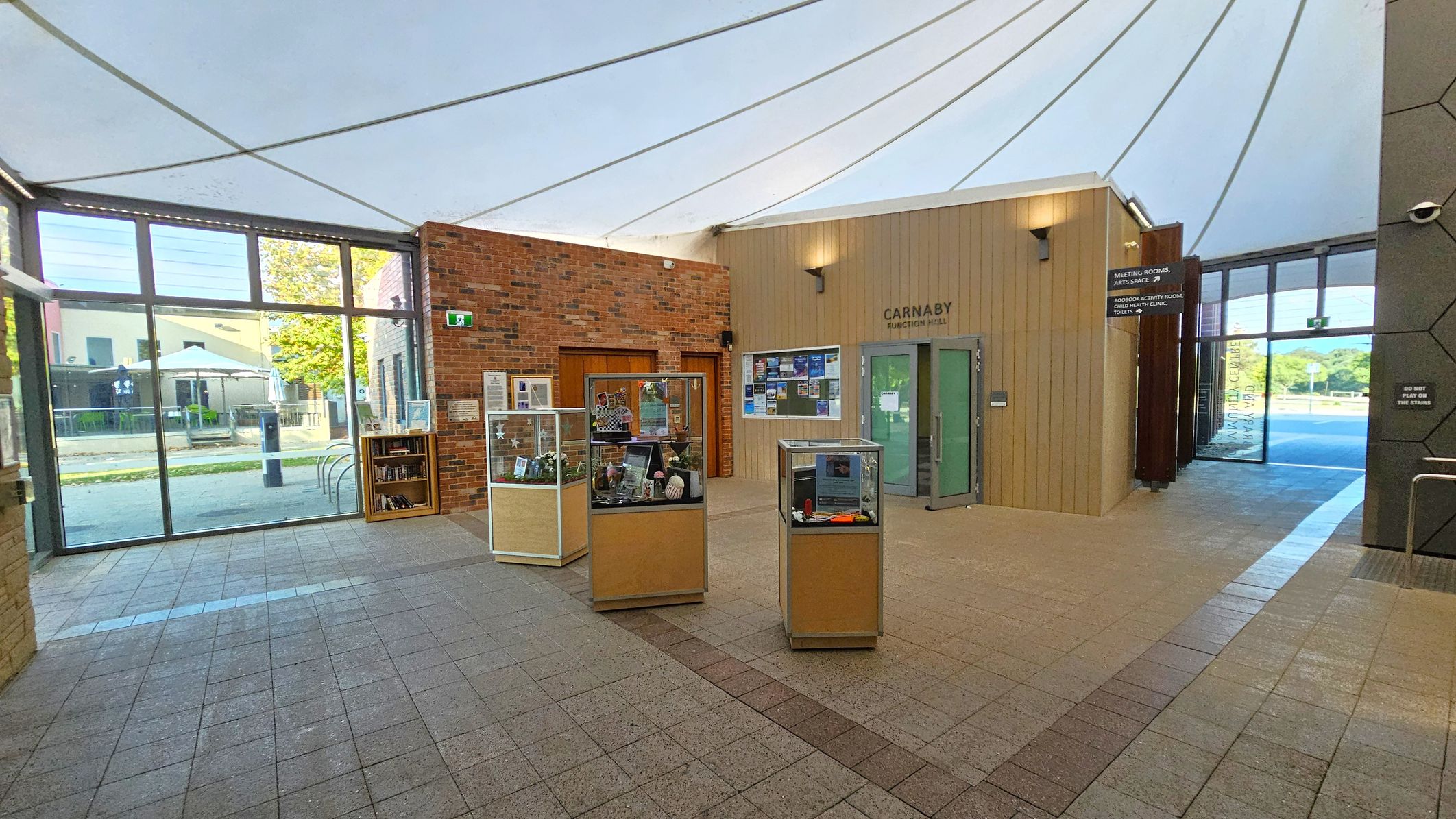 A spacious interior with a high, white tent-like ceiling. The floor is tiled in a light brown pattern. On the left, large glass windows reveal an outdoor area with trees and a building. In the centre, there are display cases with various items. The back wall is a mix of brick and wood panelling, featuring a door labelled "Carnaby Function Hall" and a bulletin board. On the right, a corridor leads to more rooms, with signs indicating meeting rooms and an arts space.