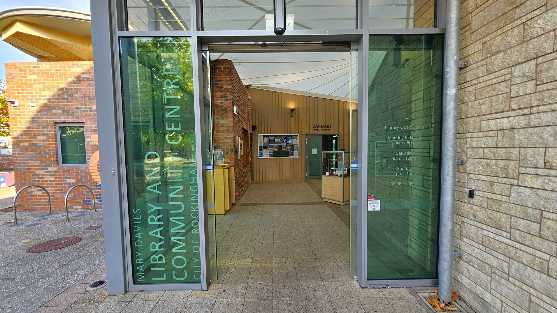 Entrance to a building with glass doors. The left door has text reading "Mary Davies Library and Community Centre, City of Rockingham." The right door displays library opening hours. Inside, a hallway with brick and wood-panelled walls leads to a notice board and a door marked "Carnaby Function Room." Display cases are on either side. The exterior features brick and stone walls, with a bicycle rack visible on the left. The floor is tiled, and the ceiling inside is white with lighting.