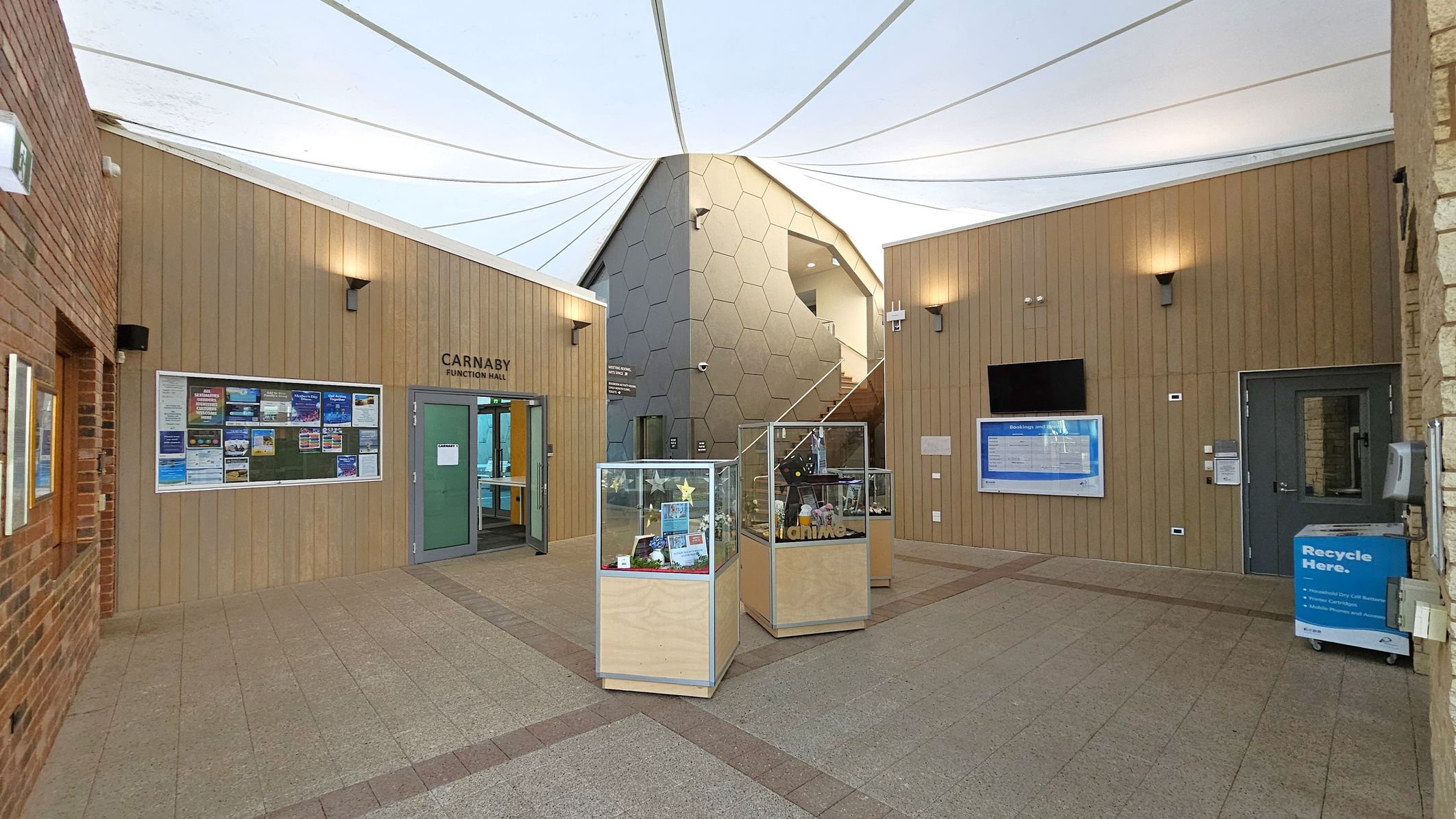 A modern indoor space with a high, white, tent-like ceiling. In the centre, a hexagonal-patterned structure with a staircase. On the left, a wooden wall with a glass door labelled "Carnaby Function Hall" and a notice board. On the right, another wooden wall with a digital screen and a recycling bin. In the foreground, three glass display cases on a tiled floor with a mix of brown and green hues.