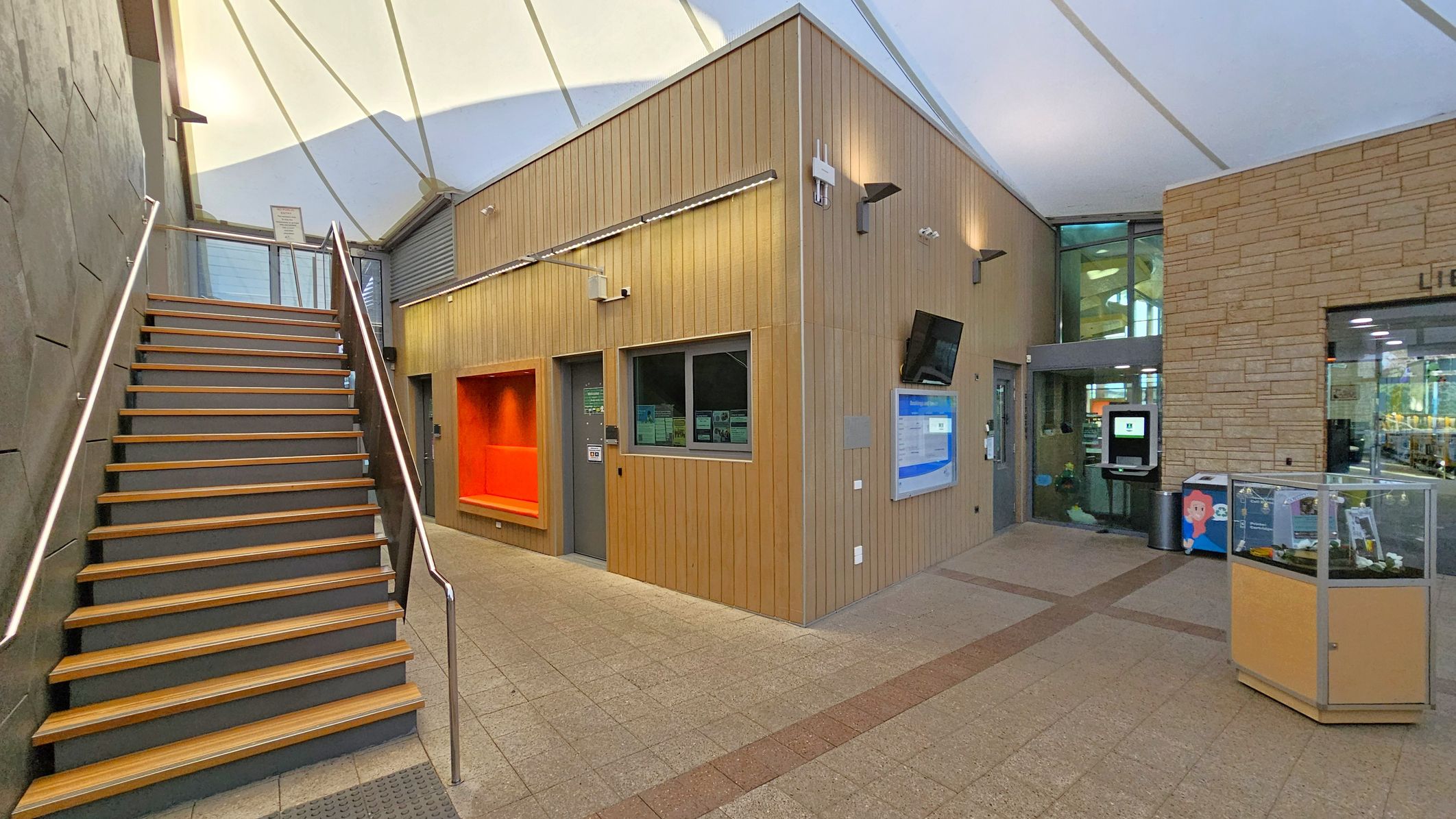 A modern interior with a staircase on the left featuring wooden steps and metal railings. The centre has a wooden-panelled wall with a recessed orange seating area, a door, and a window. A wall-mounted TV and a notice board are also present. On the right, there's a glass entrance to a library, with a display case and a digital kiosk nearby. The ceiling is white and tent-like, and the floor is tiled in a light grey pattern.