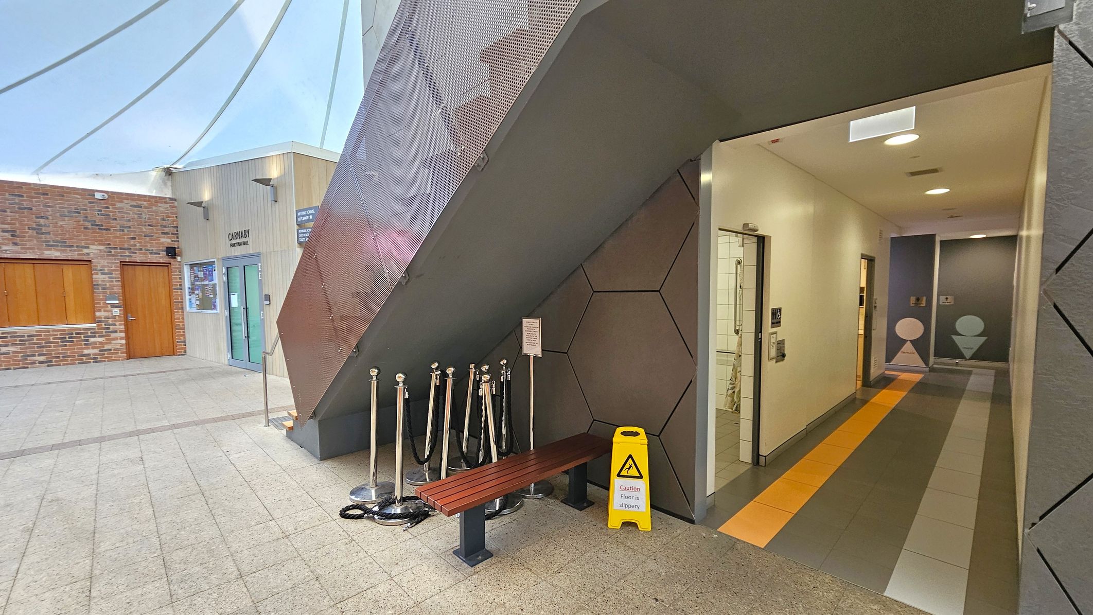 A modern indoor space with a sloped ceiling. On the left, there's a brick wall with a wooden door and a glass entrance labelled "Carnaby". In the centre, a staircase with a metal mesh railing is above a wooden bench and several stanchions. A yellow caution sign is on the floor. On the right, a corridor with tiled flooring leads to restrooms, marked by geometric symbols on the walls. The floor is tiled, and the ceiling has recessed lighting.