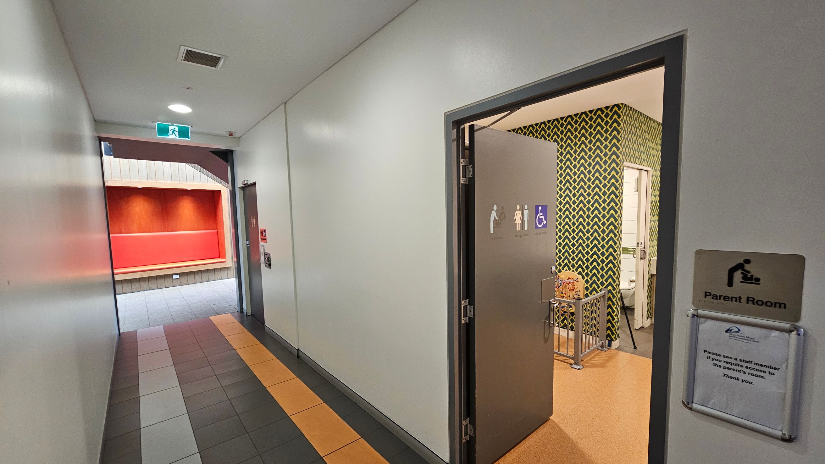 A hallway with a tiled floor featuring grey, white, and orange tiles. On the right, a door is open, leading to a parent room with a patterned wall and a baby changing station. A sign on the door indicates it is a parent room. In the background, an exit sign is visible above a doorway leading to an area with red walls and grey floor tiles. The ceiling is plain with a light fixture and a vent.