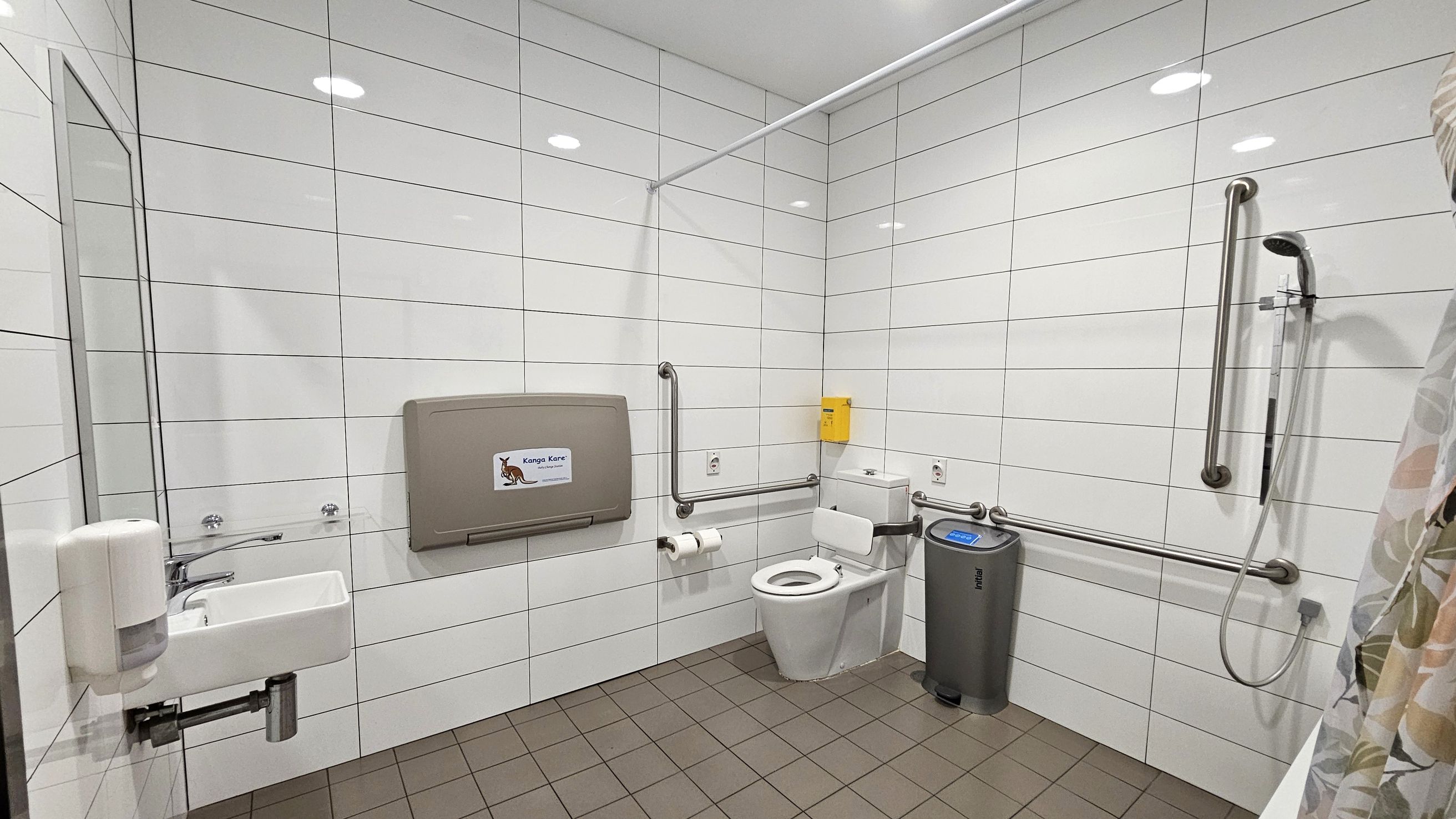A spacious, accessible bathroom with white tiled walls and a brown tiled floor. On the left, a small sink with a soap dispenser is mounted on the wall. A baby changing station is positioned above the sink. In the centre, a toilet with grab bars on both sides is visible, along with a toilet paper holder. On the right, a shower area with a handheld showerhead and additional grab bars. A grey bin is placed near the toilet. The ceiling has recessed lighting.
