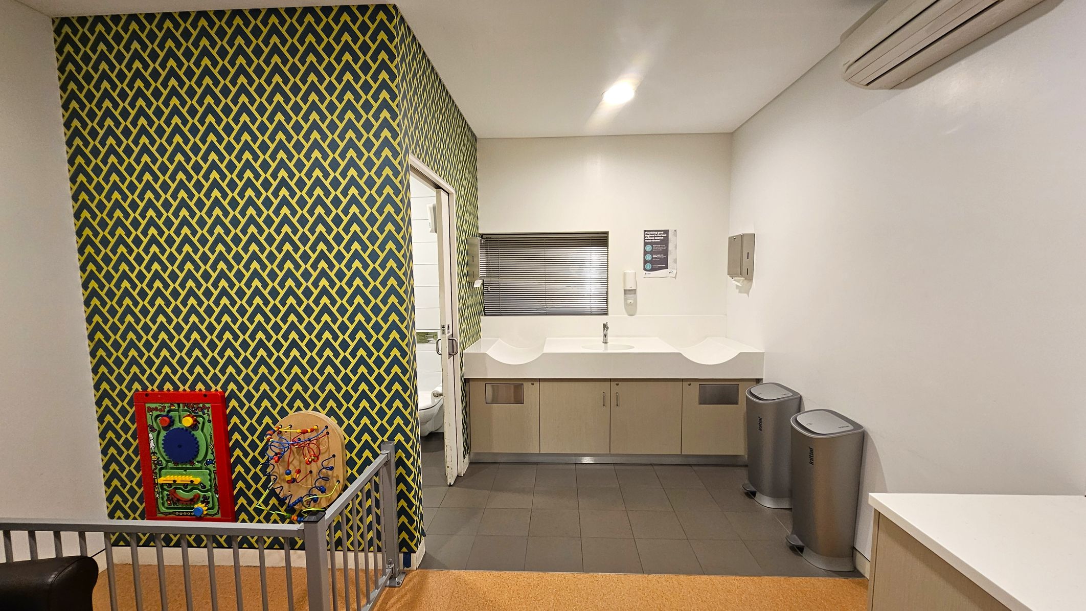 A room with a patterned wall on the left featuring a yellow and green geometric design. Two interactive children's wall toys are mounted on this wall. A small gate is visible in the foreground. In the centre, there is a white sink with a tap, set against a plain wall with a small window. Two silver bins are placed on the right side. The floor is tiled in grey, and the ceiling is plain white with a light fixture. An air conditioning unit is mounted on the right wall.