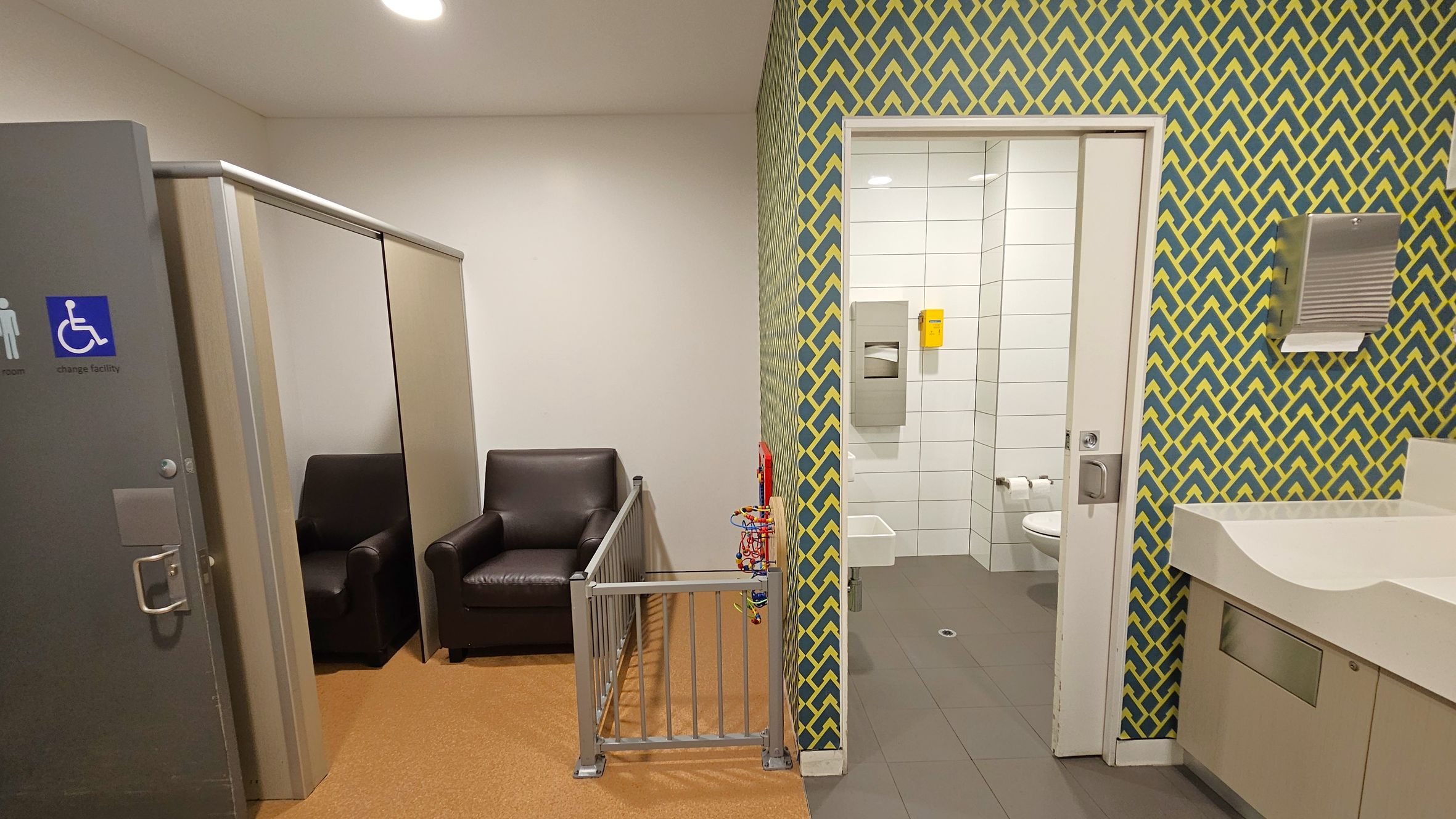 A room with a grey door on the left featuring a wheelchair-accessible sign. Inside, there are two dark leather chairs and a small play area with a metal gate and a bead maze toy. The wall has a green and yellow geometric pattern. On the right, an open door leads to a tiled bathroom with a toilet, paper dispenser, and a small sink. A white countertop with a paper towel dispenser is visible on the far right. The floor is a mix of brown and grey tiles.