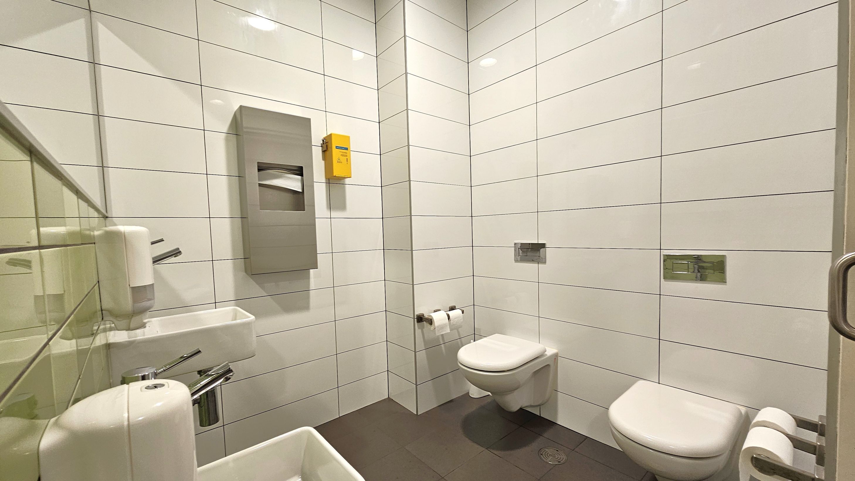 A clean public restroom with white tiled walls and a dark tiled floor. On the left, there is a small white sink with a soap dispenser above it. In the centre, a wall-mounted toilet paper dispenser and a yellow box are visible. On the right, two wall-mounted toilets are installed side by side, each with a toilet paper holder. The ceiling has recessed lighting, and a metal door handle is visible on the right edge.