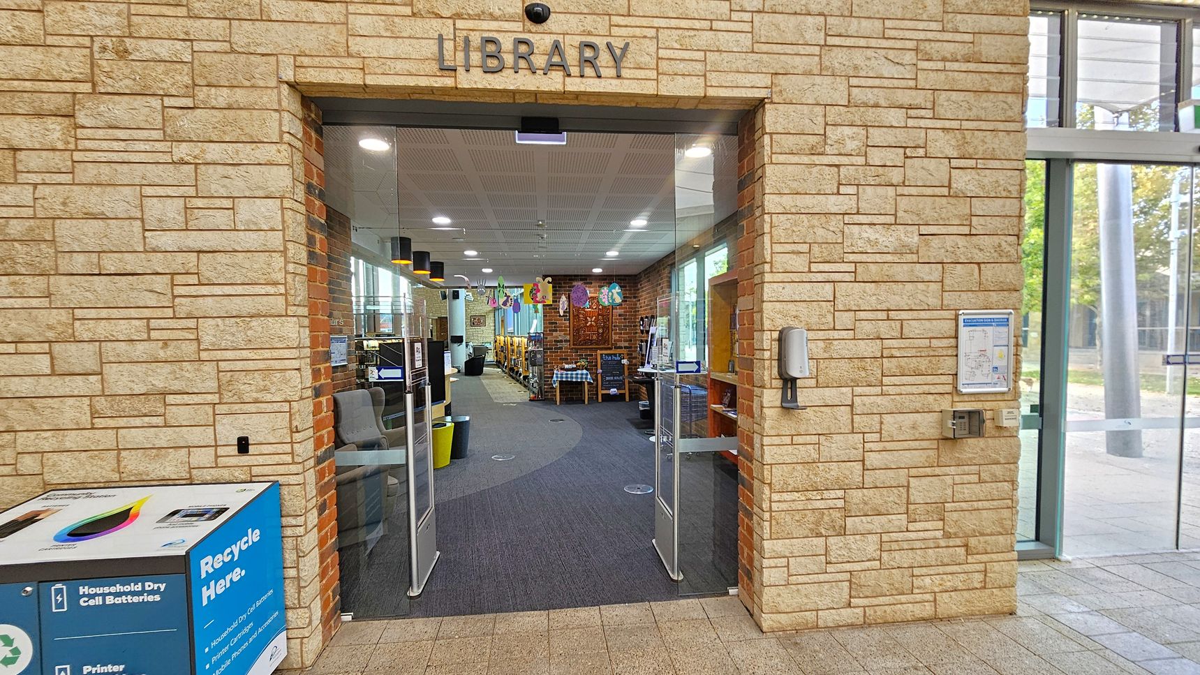 Entrance to a library with a stone brick facade. The word "LIBRARY" is displayed above the glass doors. Inside, there are bookshelves and a seating area visible. A recycling bin is on the left, labelled for household dry cell batteries and printer cartridges. On the right, a hand sanitiser dispenser and a notice board are mounted on the wall. The floor is tiled outside and carpeted inside. Ceiling lights illuminate the interior.