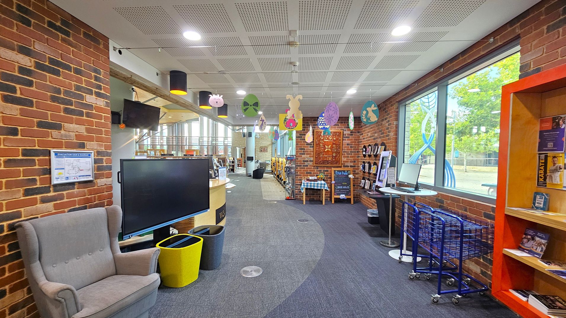 A library interior with a brick wall on the left featuring a grey armchair and a large screen. A yellow bin is nearby. The ceiling has recessed lighting and hanging decorations. On the right, a large window shows a view of trees and a blue sculpture outside. Below the window, there are blue trolleys and a small round table with a computer. An orange bookshelf displays various books and pamphlets. The floor is carpeted in grey.
