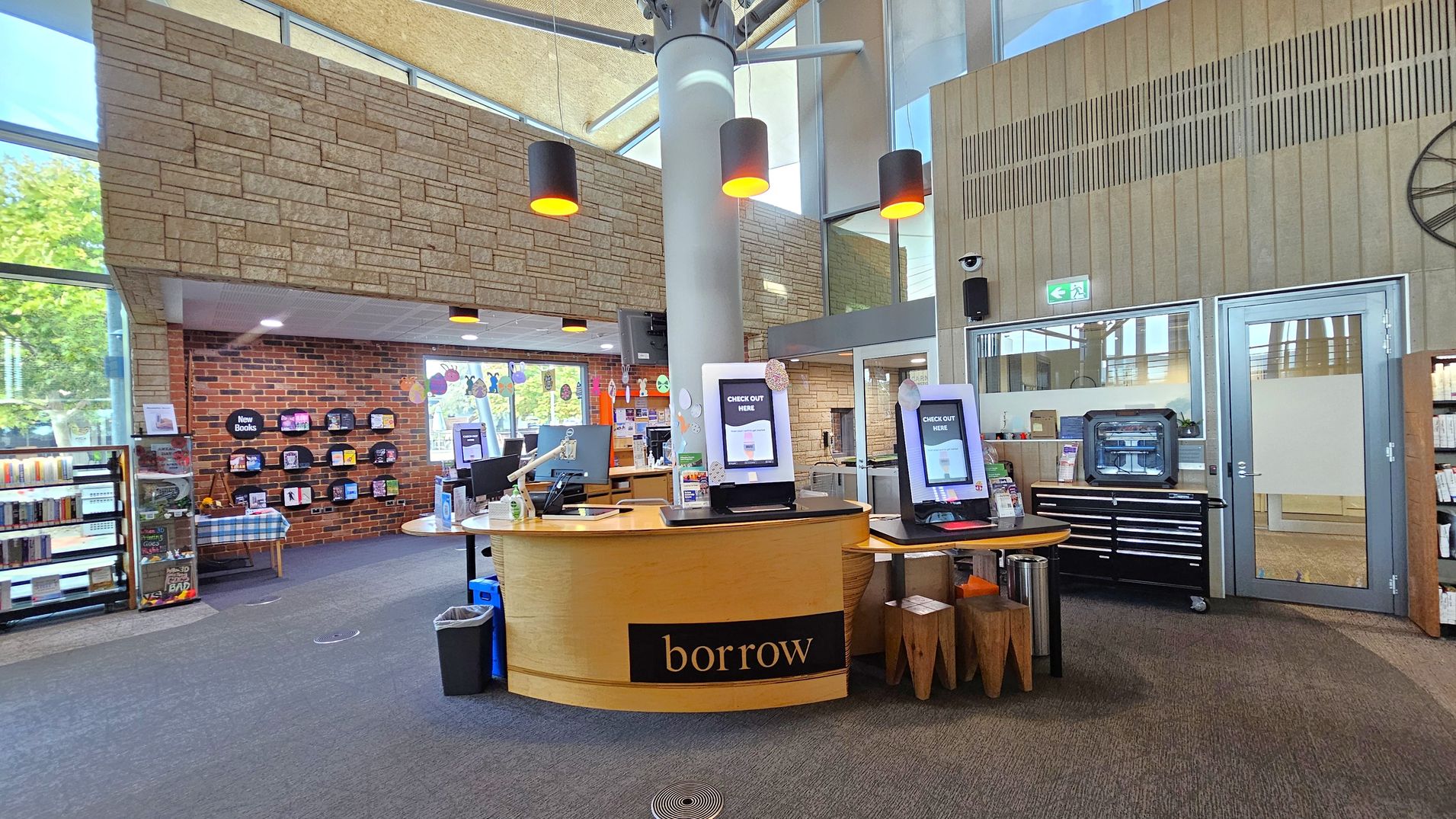 A library interior with a central circular wooden desk labelled "borrow" featuring self-checkout screens. To the left, bookshelves and a brick wall with a "New Books" display. The right side has a glass door and a black cabinet with drawers. The ceiling is high with large windows and hanging lights. The floor is carpeted in grey tones.
