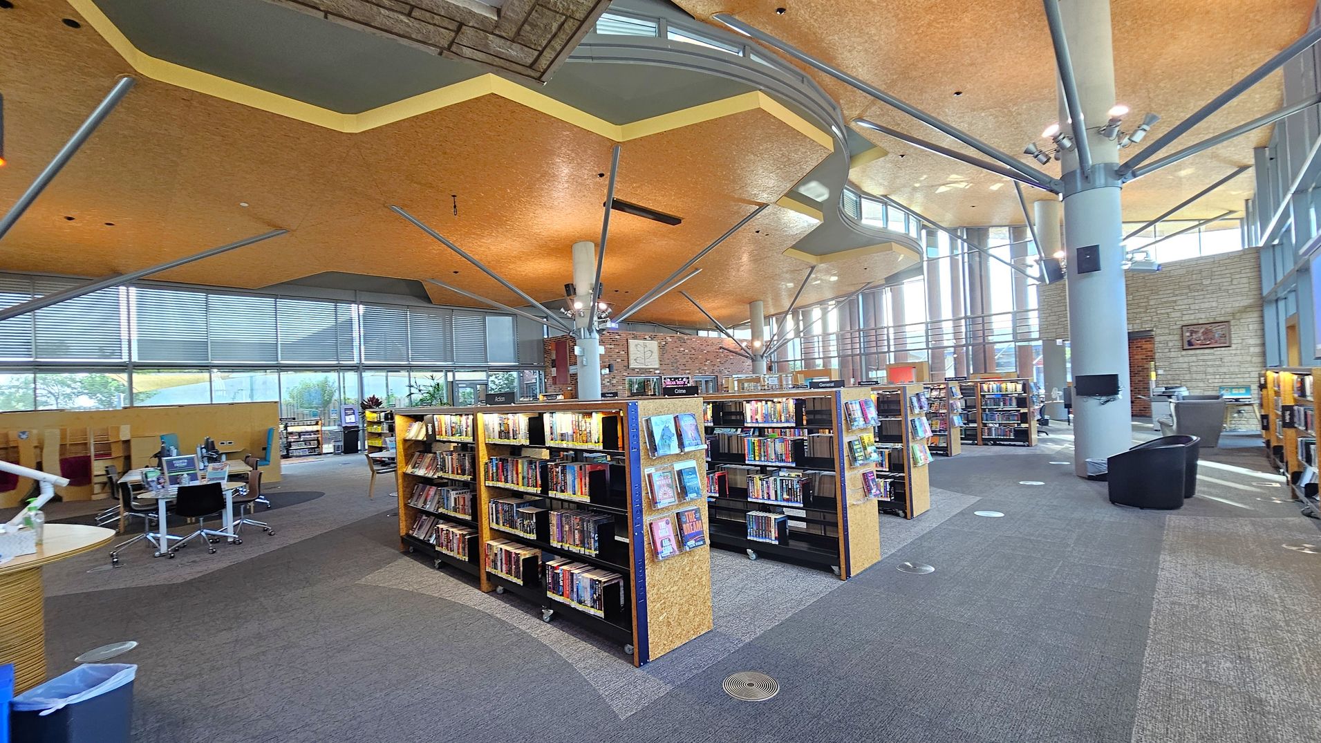 A spacious library interior with several bookshelves filled with books in the centre. On the left, there are tables with chairs and computers. The right side features more bookshelves and a seating area with armchairs. The ceiling is uniquely designed with geometric patterns and supported by metal beams. Large windows line the background, allowing natural light to illuminate the space. The floor is carpeted in a mix of grey and purple tones.