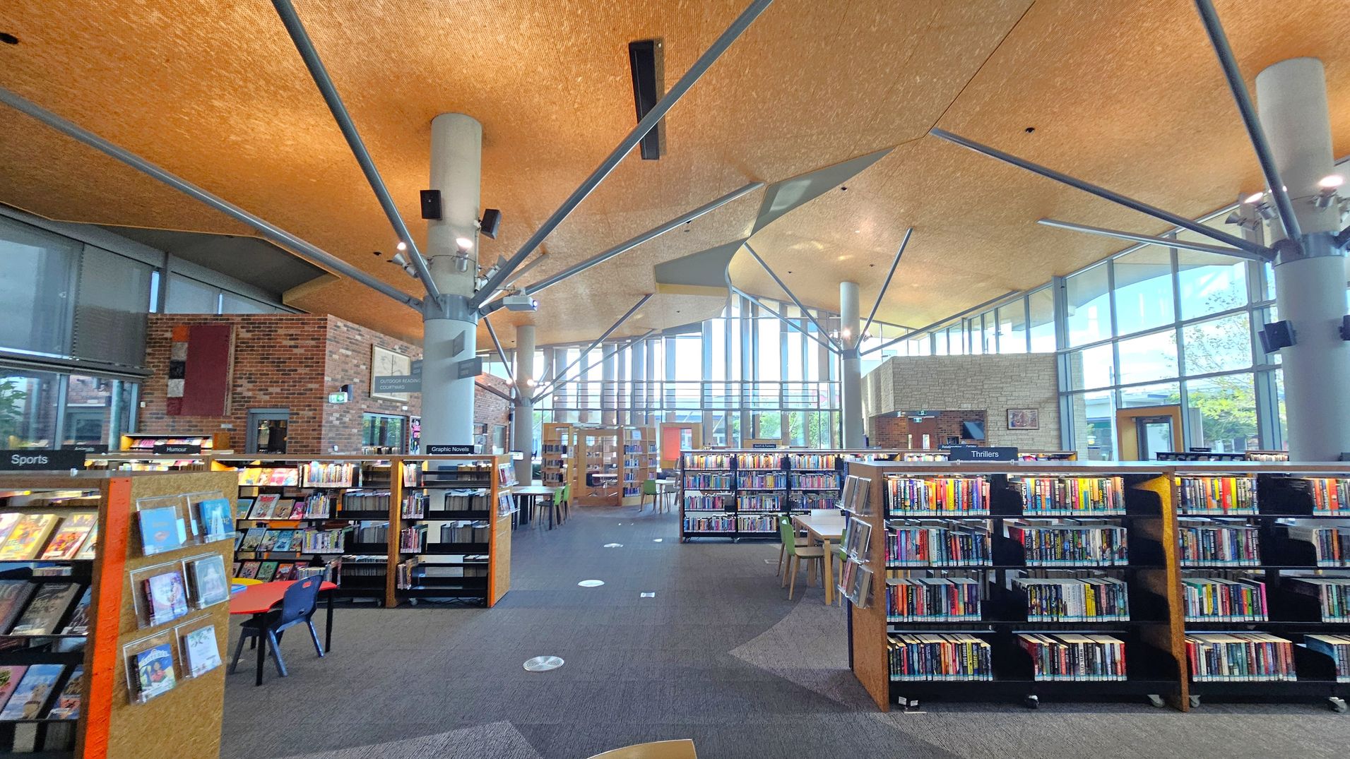 A modern library interior with a high, angular ceiling featuring exposed beams. In the foreground, bookshelves are labelled with categories like "Sports," "Humour," "Graphic Novels," and "Thrillers." A small table with chairs is visible on the left. The floor is carpeted in a mix of grey and purple tones. Large windows in the background allow natural light to flood the space, and brick walls add texture. Pillars support the ceiling, and there are tables and chairs for reading.