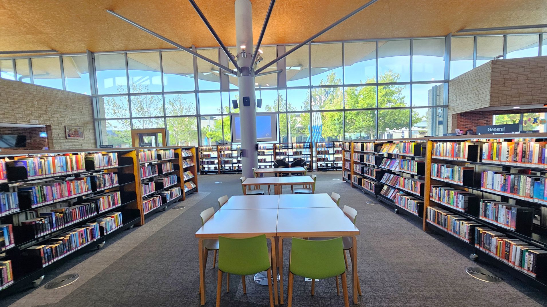 A library interior with a high ceiling and large windows in the background, allowing natural light to illuminate the space. In the foreground, there are tables with green and white chairs. Bookshelves filled with books line both the left and right sides. A central pillar supports the ceiling, and the floor is carpeted. The windows offer a view of trees outside.