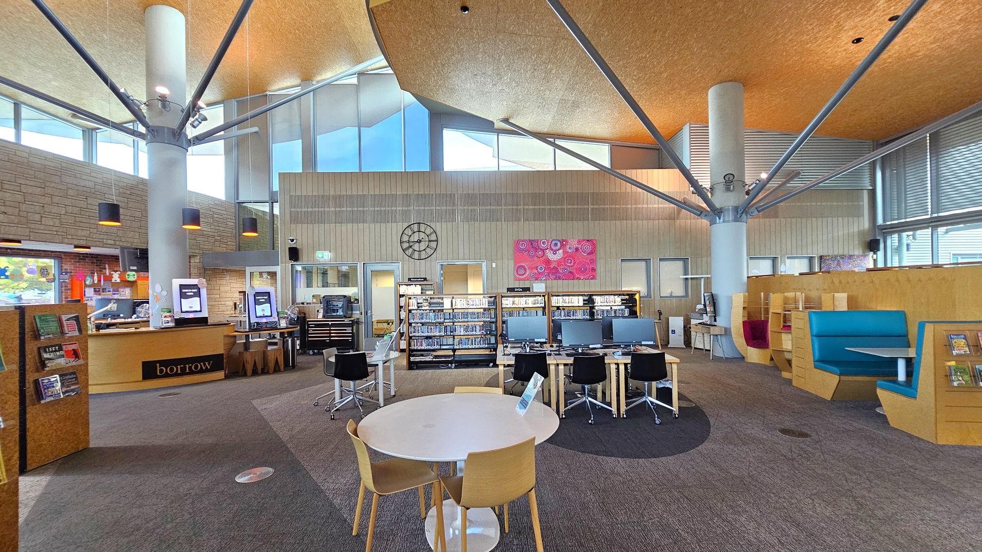 A modern library interior with a high, angular ceiling and large windows. In the foreground, a round table with wooden chairs is on a carpeted floor. To the left, a counter with a "borrow" sign and self-service machines. In the centre, rows of bookshelves and computer stations with black chairs. The right side features colourful seating booths. A large clock and a vibrant artwork are mounted on the back wall. Structural columns with metal beams are visible throughout.