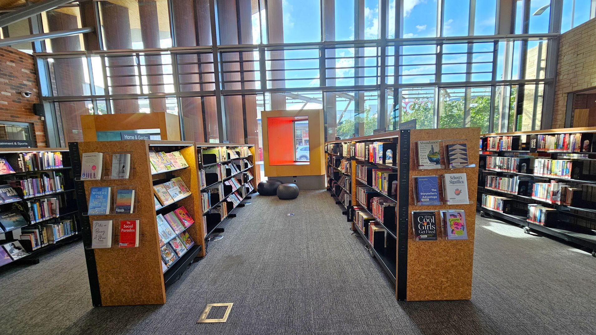 Bookshelves line both sides of a library aisle, filled with various books and magazines. The shelves are labelled with categories like "Home & Garden". In the centre, a large window with vertical metal bars allows natural light to flood in, and a bright orange window seat is visible. Two black bean bags are placed on the grey carpeted floor. The ceiling is high with exposed beams, and the walls are a mix of brick and glass.