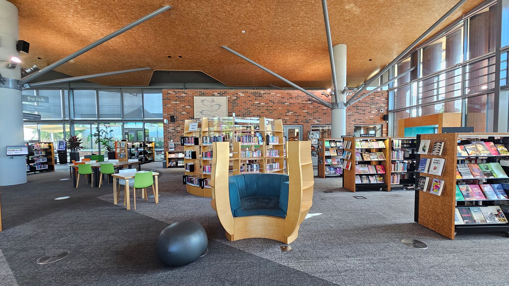 A modern library interior with a high, textured ceiling supported by metal beams. In the centre, there is a unique, semi-circular wooden chair with blue cushioning. Surrounding it are curved bookshelves filled with books. On the left, there are tables with colourful chairs and a grey exercise ball. The right side features more bookshelves with various books on display. The background has large windows and a brick wall with a decorative panel. The floor is carpeted in shades of grey and purple.
