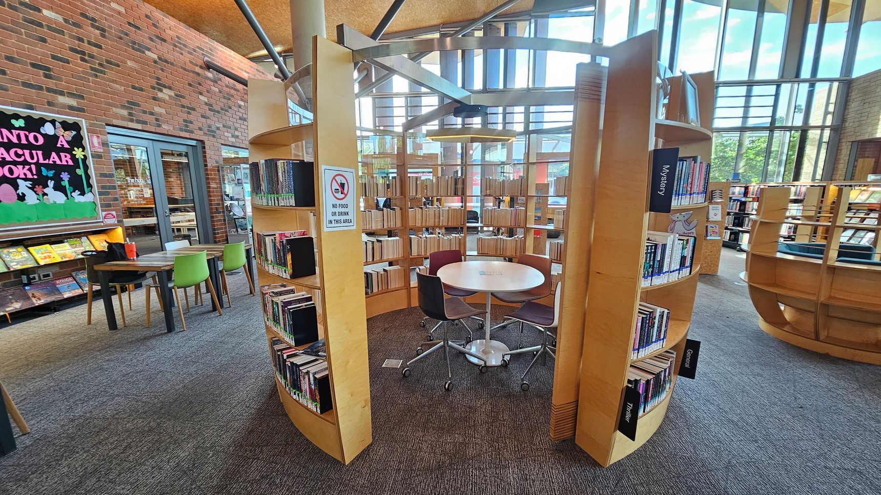 Circular wooden bookshelves form an enclosed reading area with a round table and chairs in the centre. A sign on the shelves reads "No food or drink in this area." To the left, a brick wall features a colourful bulletin board and a table with green chairs. Large windows in the background allow natural light to fill the space. The carpet is grey with purple accents.