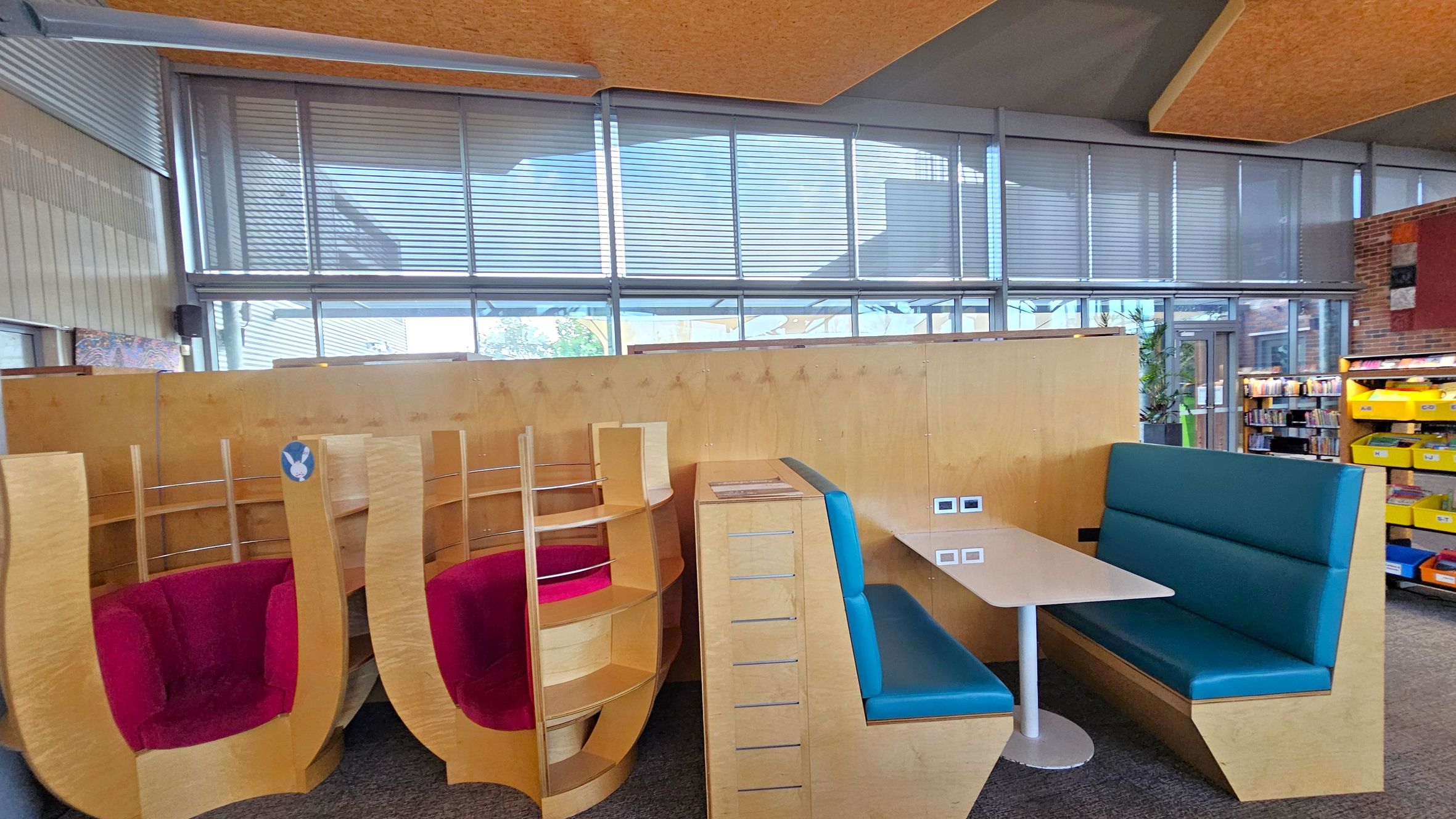 On the left, there are two wooden pod-like chairs with red cushions, featuring curved backs and integrated shelving. In the centre, a wooden partition separates the seating areas. On the right, a booth with blue cushioned seating surrounds a white table. The background shows large windows with blinds, and a glimpse of bookshelves with yellow bins. The ceiling has a textured, wooden appearance, and the floor is carpeted.