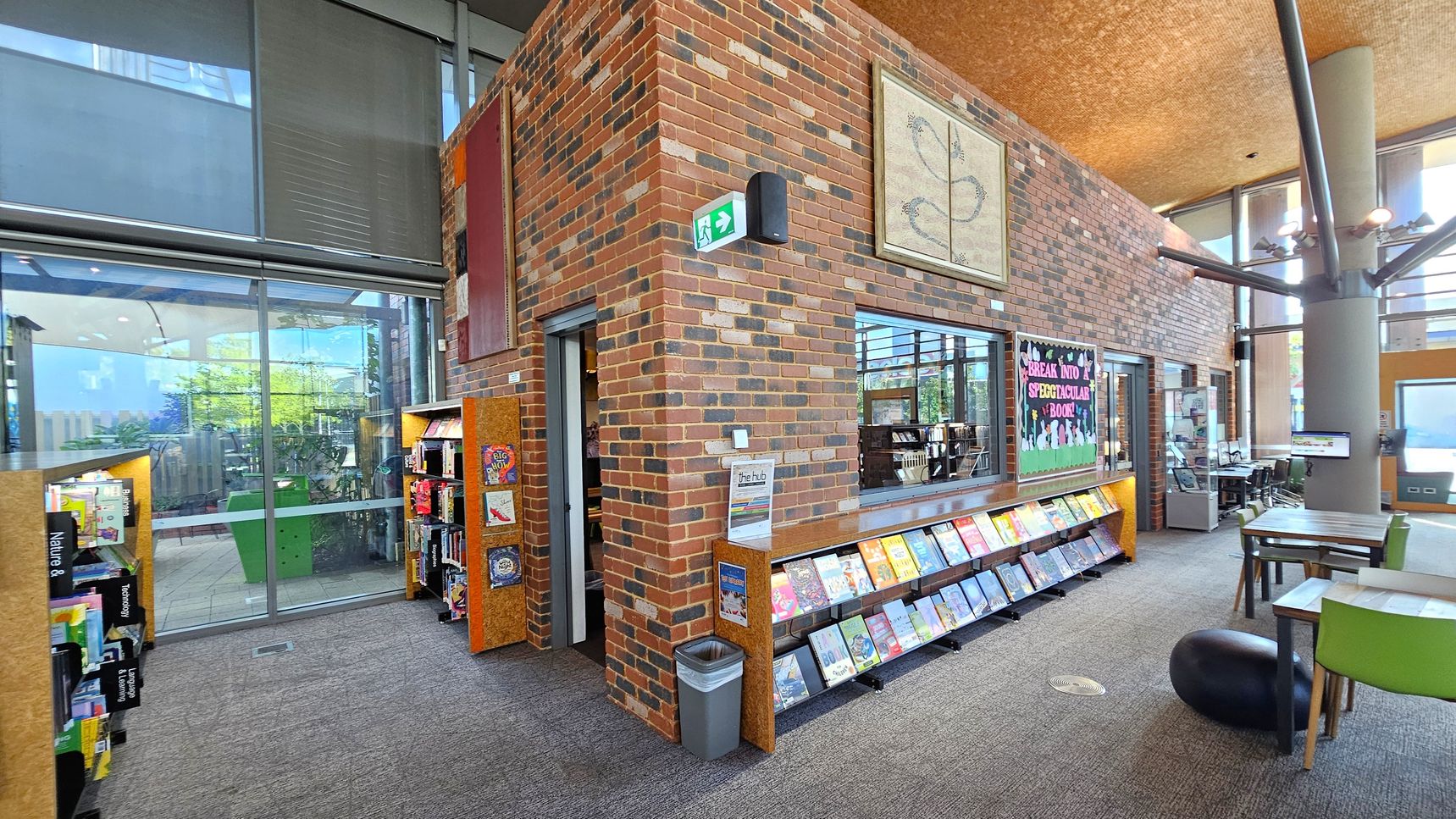 A library interior with a brick wall in the centre. On the left, there are bookshelves filled with books, labelled with categories like "Nature" and "Transport". A large window with blinds is behind the shelves. On the right, a display of books is set against the wall, with a colourful sign above. A table with green chairs and a black stool is nearby. The ceiling is textured, and a column with light fixtures is visible. The floor is carpeted, and a small bin is near the wall.