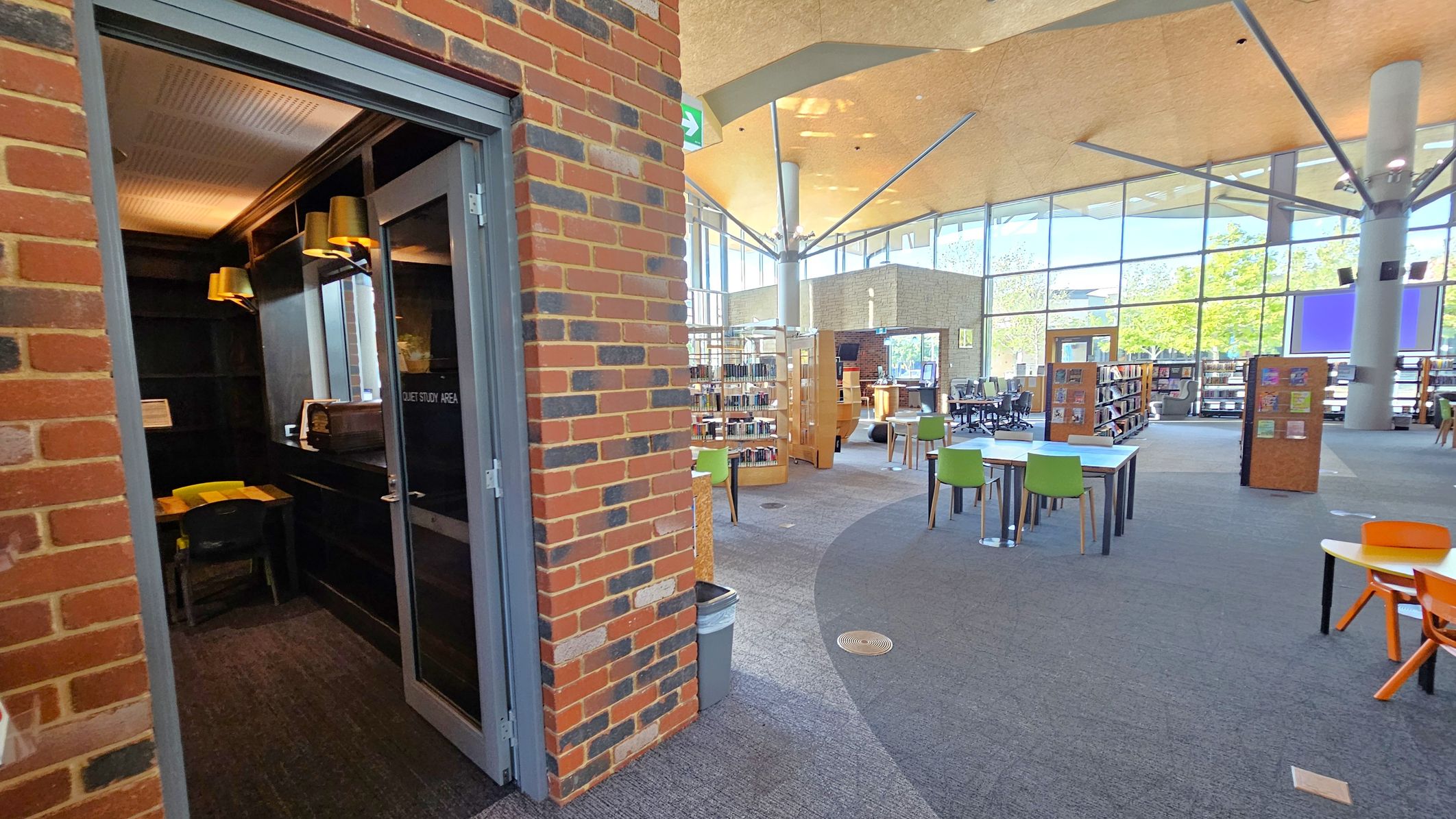 On the left, a small room with a sign reading "Quiet Study Area" features a desk, chair, and warm lighting. The room is enclosed by a brick wall. On the right, an open library space has tables with green and orange chairs, bookshelves, and large windows allowing natural light. The ceiling is high with exposed beams, and the floor is carpeted in a mix of grey and purple tones. A rubbish bin is near the entrance to the study area.
